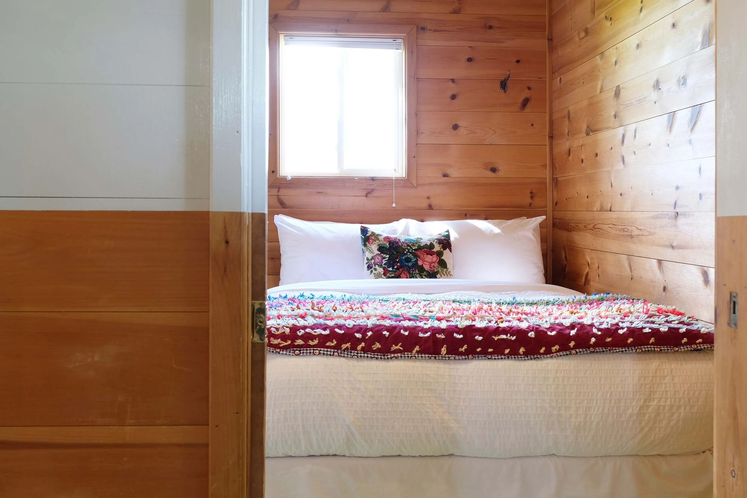 A cozy bedroom with wood-paneled walls, a window allowing natural light, a bed with white pillows, a colorful floral pillow, and a multicolored blanket.