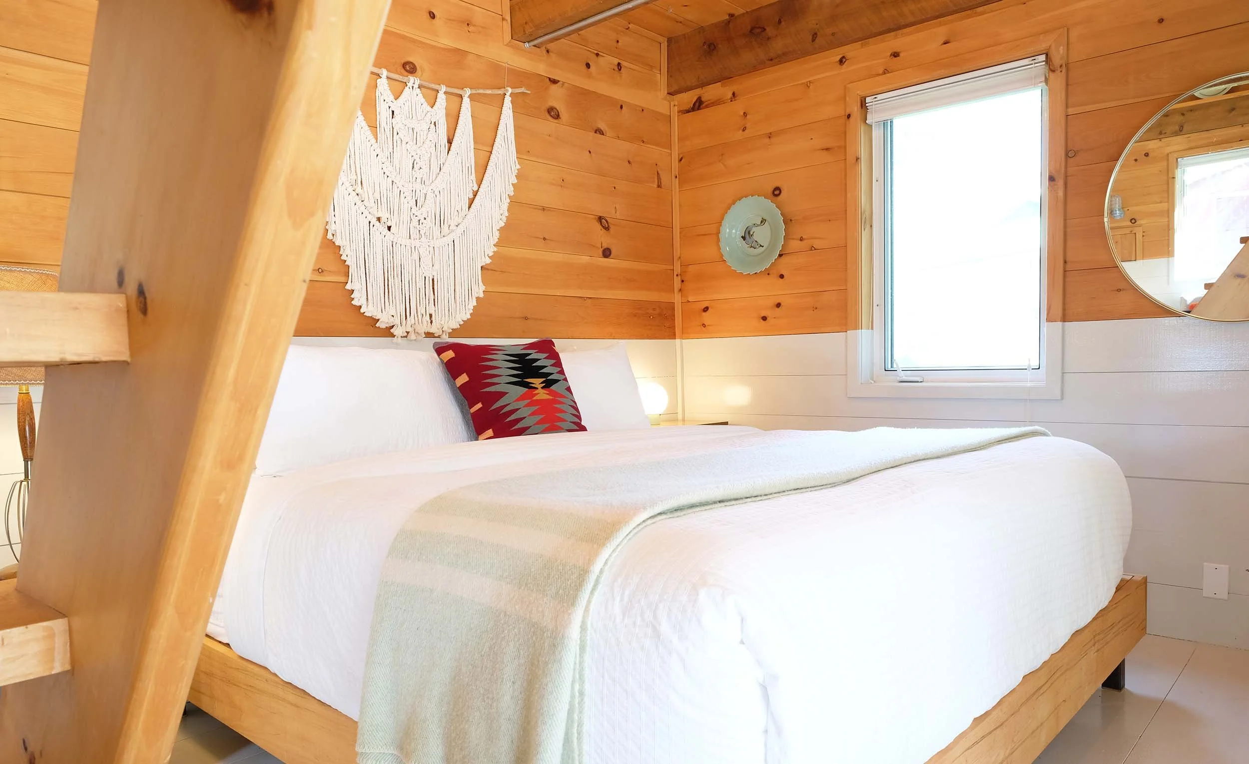 A bedroom with wood-paneled walls and ceiling, featuring a large bed with white bedding and a colorful geometric pillow. There's a macramé wall hanging, a decorative plate, a round mirror, and a window with blinds.