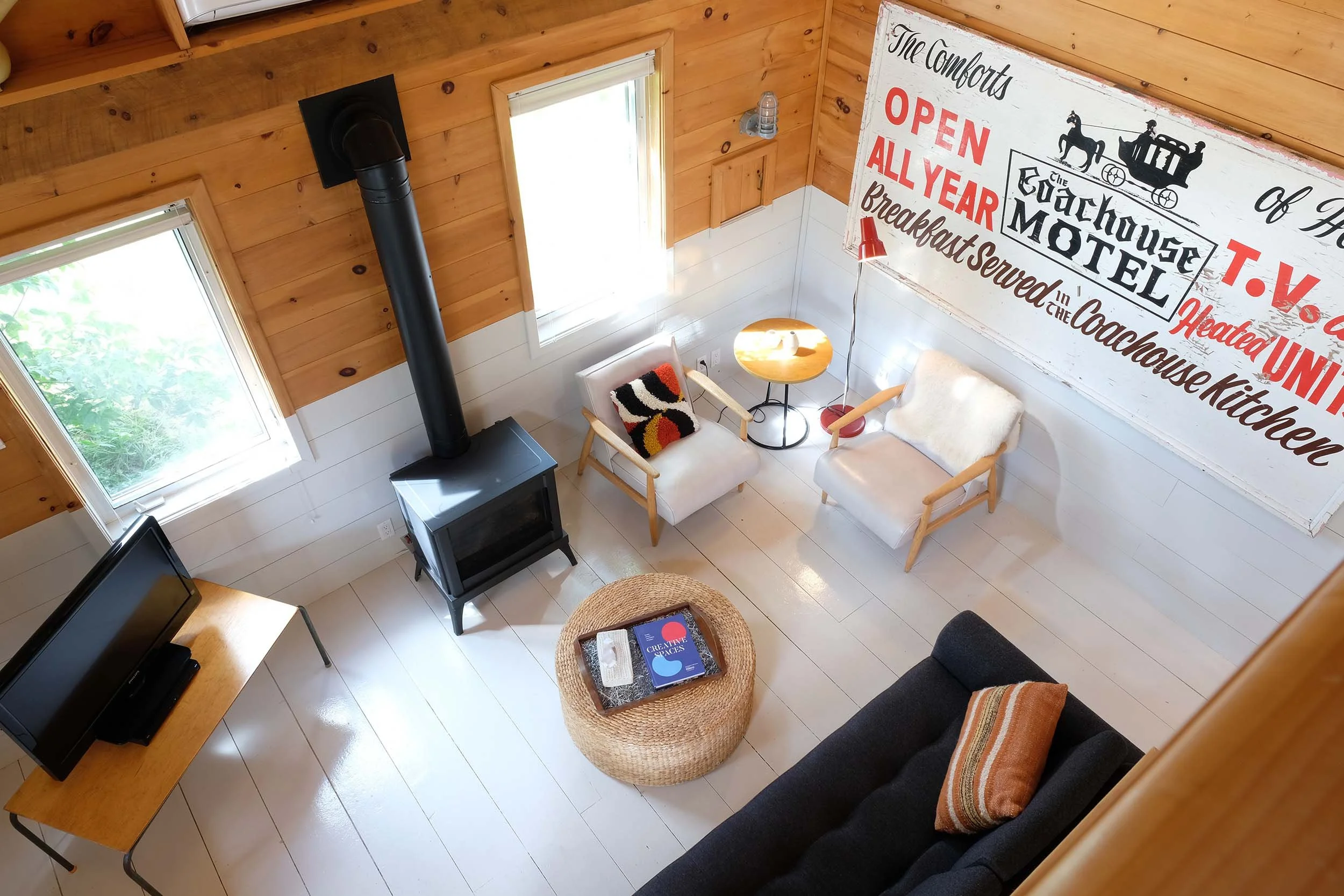 A cozy living room with wood-paneled walls and white floors, featuring a black wood stove, two armchairs with cushions, a small round wooden table, a wicker ottoman with books on it, and a large wall sign with vintage train artwork and text about an all-year breakfast service.