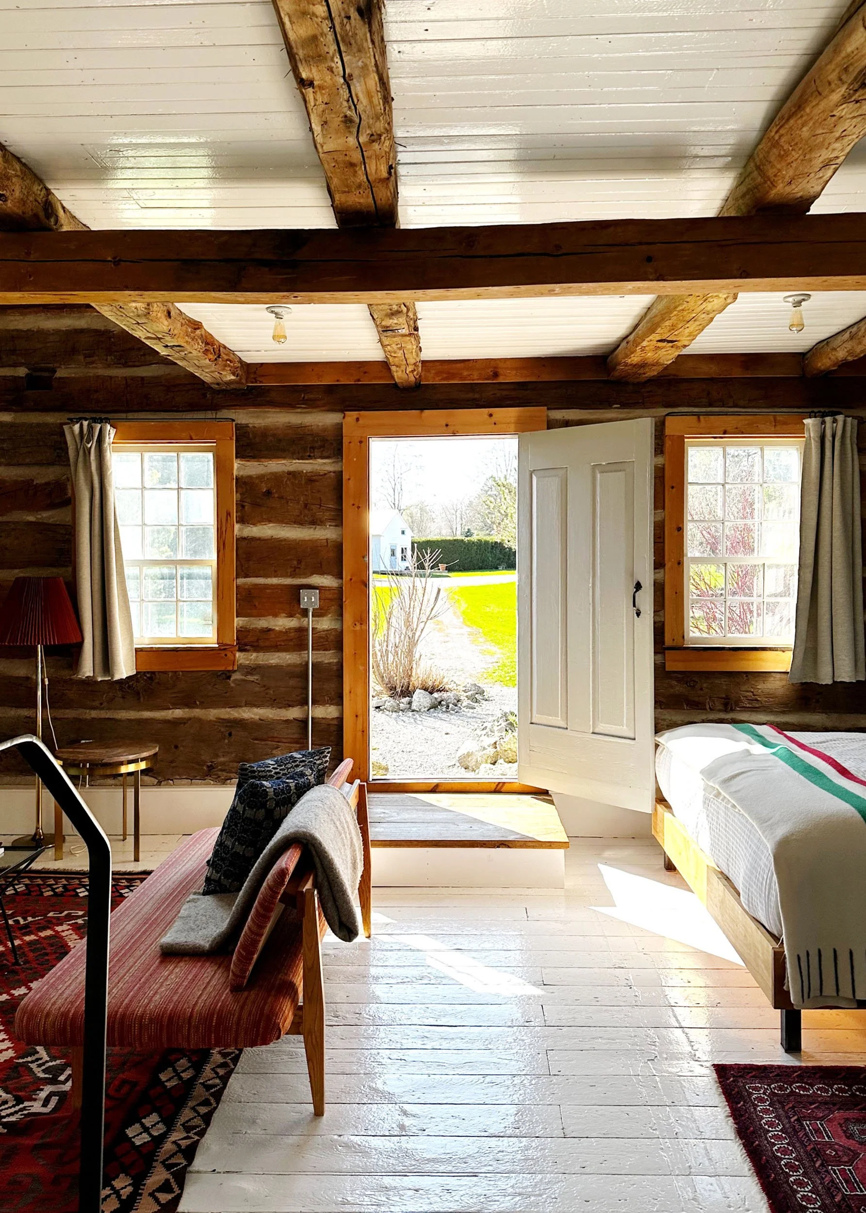 Interior of a rustic wooden cabin with white painted wooden floors, exposed wooden beams, and a white ceiling. There are two windows with cream curtains, an open door leading outside showing a house and greenery, a bed with a white blanket and colorful stripes, some chairs, a red patterned rug, and various household items.