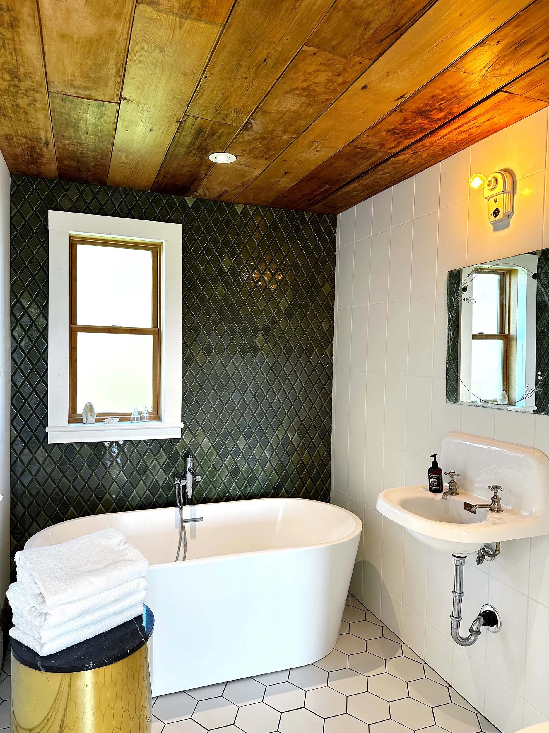 Bathroom with a white bathtub, a black and gold side table with towels, a small sink with a bar soap, a mirror, green and white tiled walls, and a wooden ceiling with a window.