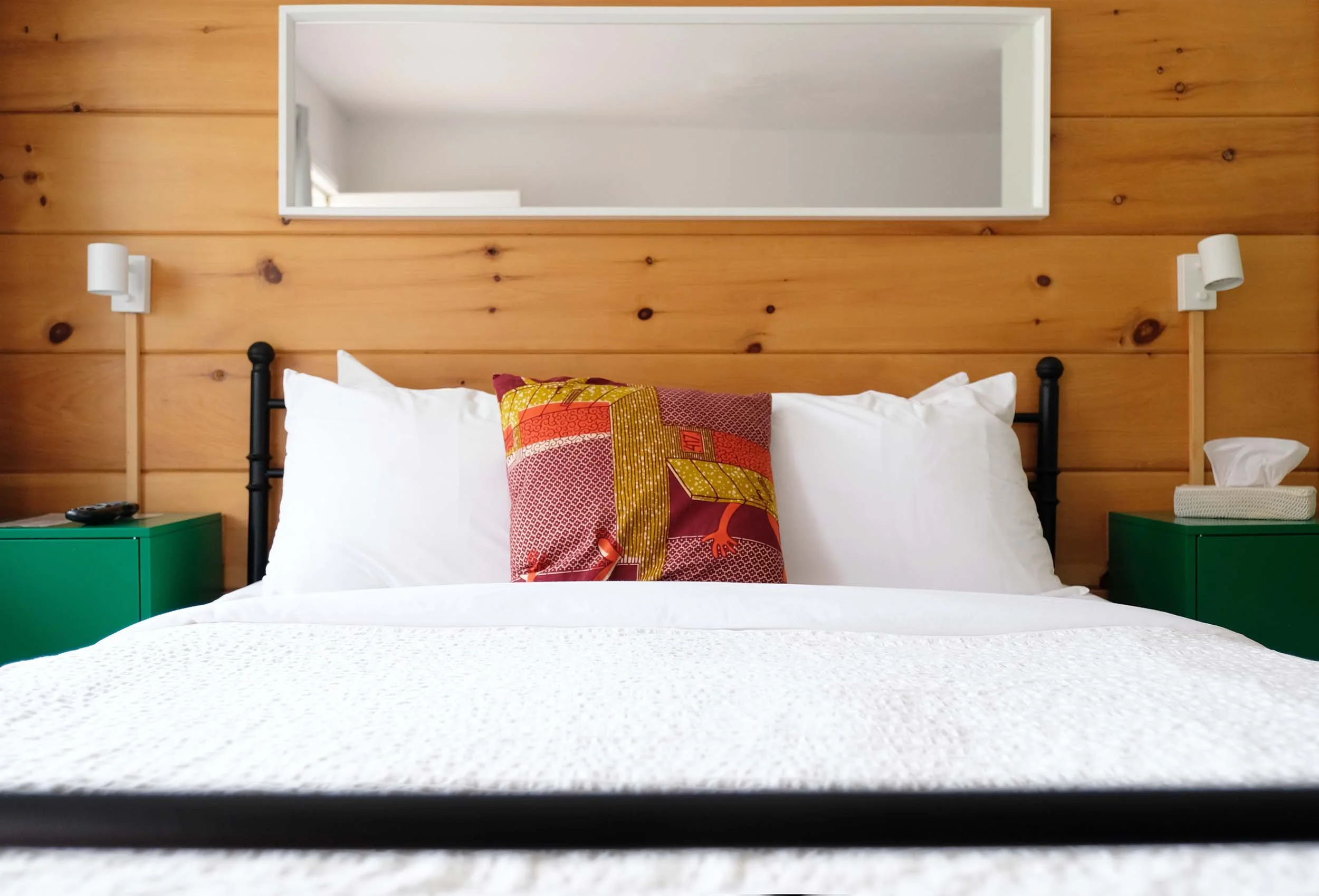 Photo of a bedroom with a wooden wall, a mirror, and bed with white bedding and a colorful pillow. Two white wall-mounted lamps on either side of the mirror, and green nightstands.