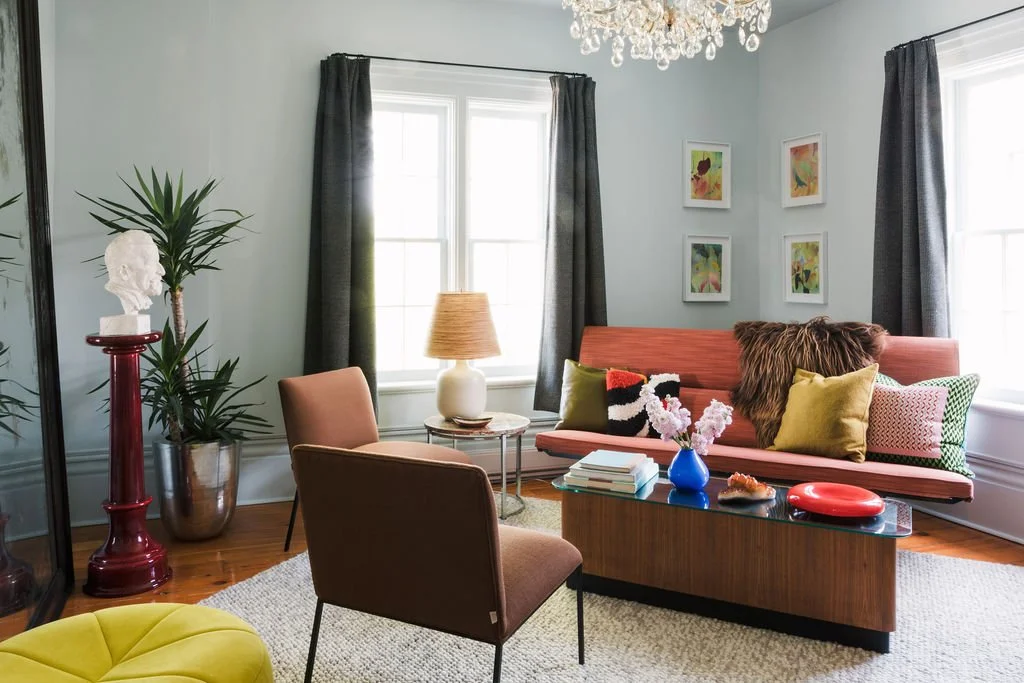 Living room with a pink sofa, colorful pillows, a wooden coffee table with decor items, a pink armchair, a table with a lamp, a tall plant, and framed artwork on the walls.
