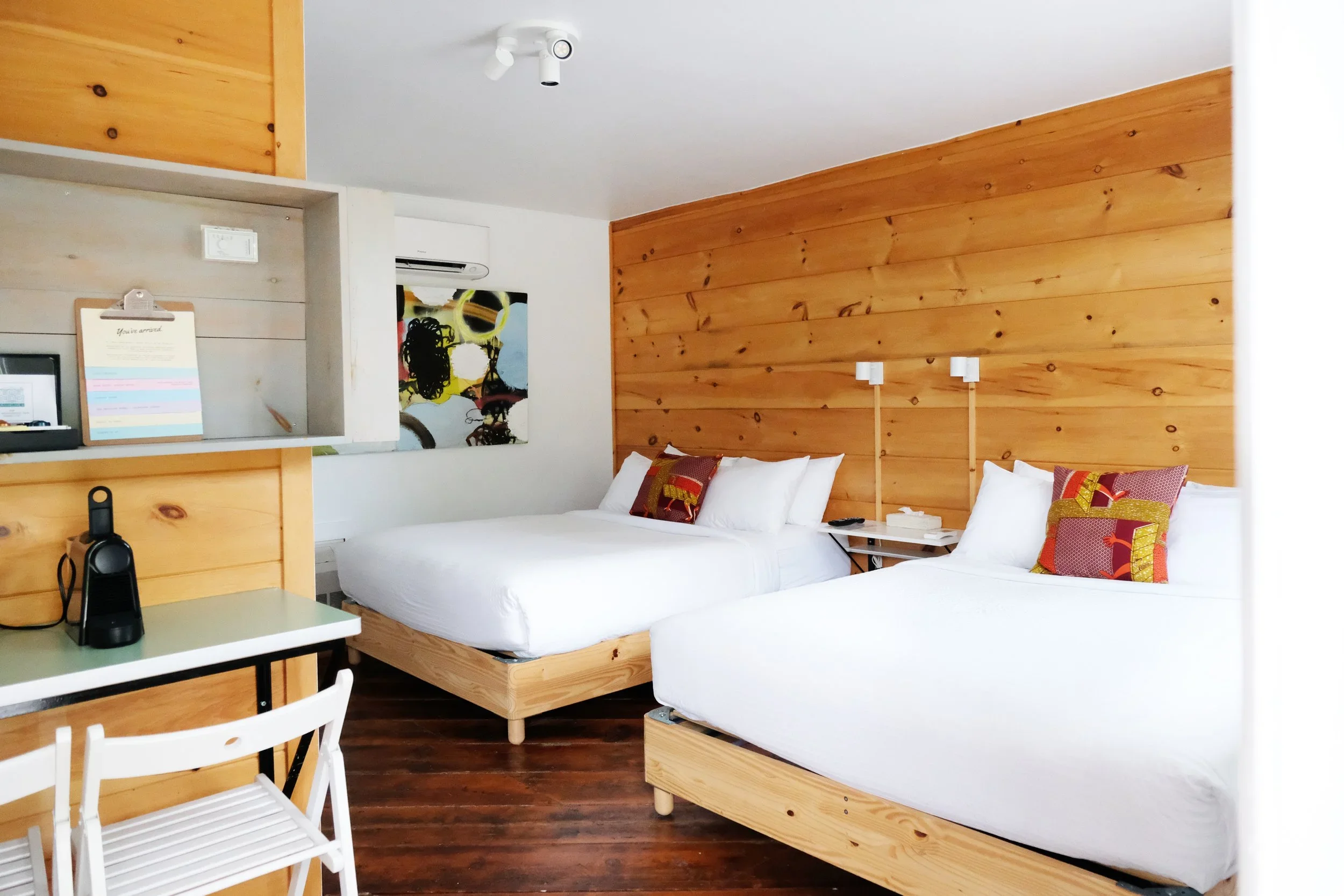 Hotel room with two beds, wooden headboard wall, colorful pillows, and a small white table with chairs and a black device.