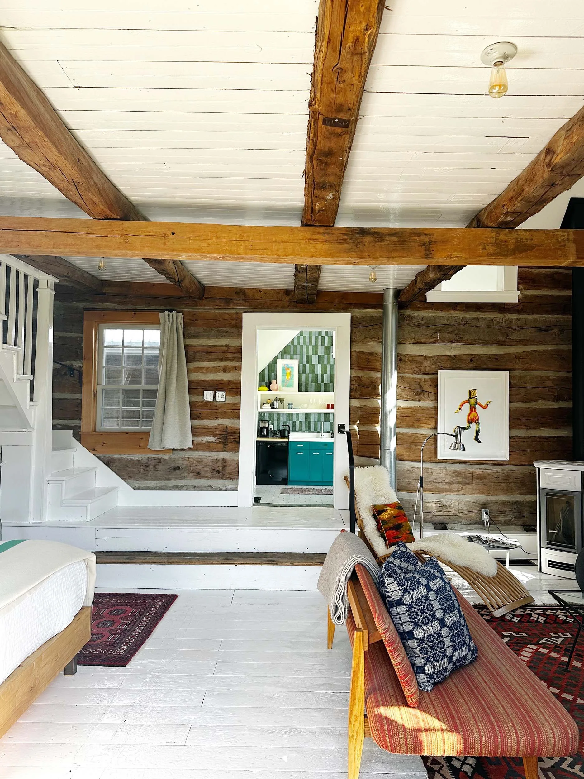 Interior of a rustic living space with white painted floorboards, wooden beams on the ceiling, and wood-paneled walls. There is a beige couch, a wooden chair with a plaid cushion and blankets, and a colorful geometric rug. A window with beige curtains allows natural light to enter. The room also features artwork on the wall, a small kitchenette with teal cabinets, and various decorative items.