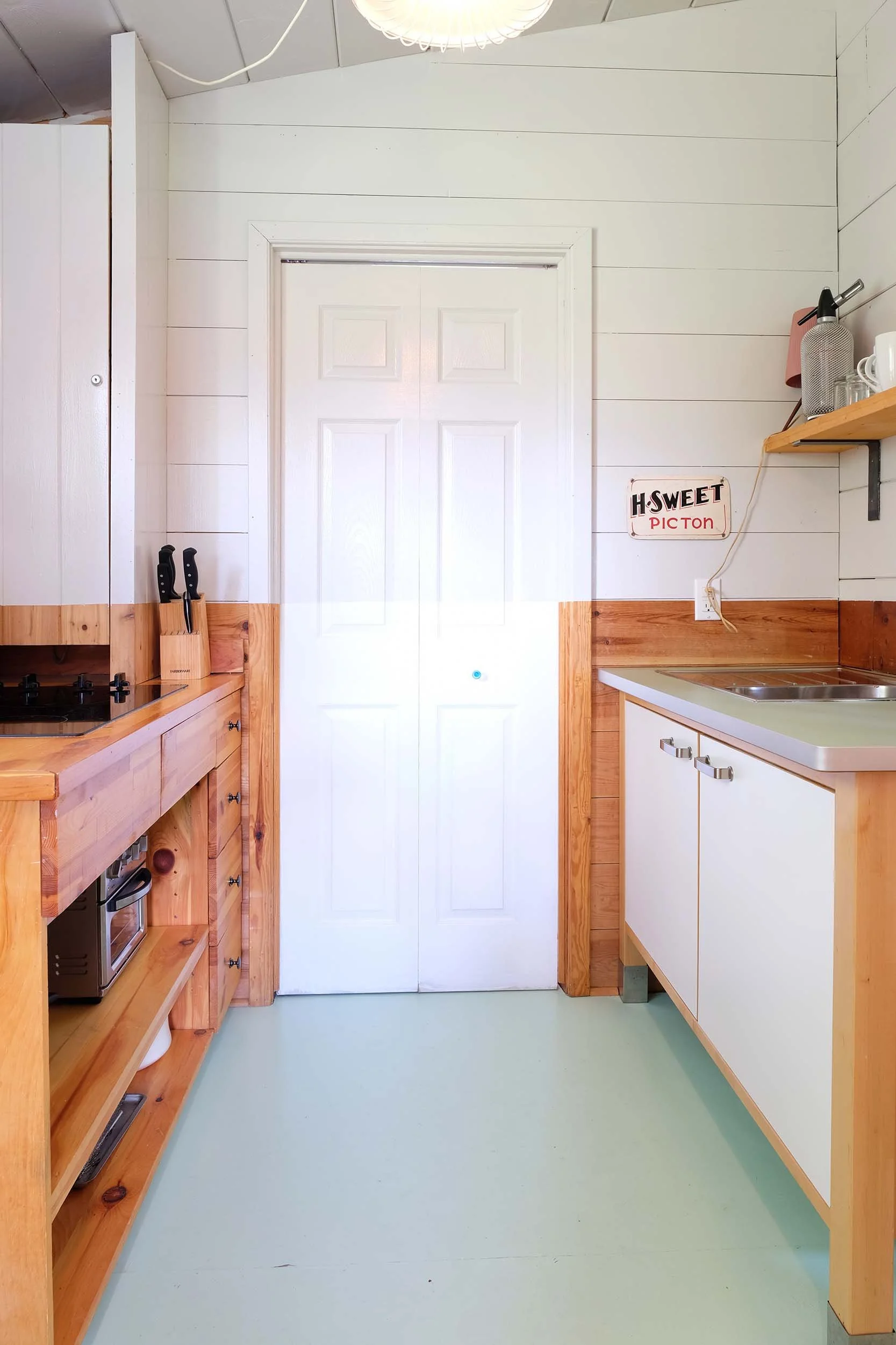 A small kitchen with white wall paneling, wooden trim, and light green flooring. On the left, there is a wooden counter with a toaster oven and two knives in a knife block. On the right, there is a sink with white cabinets below, a shelf with cups and utensils