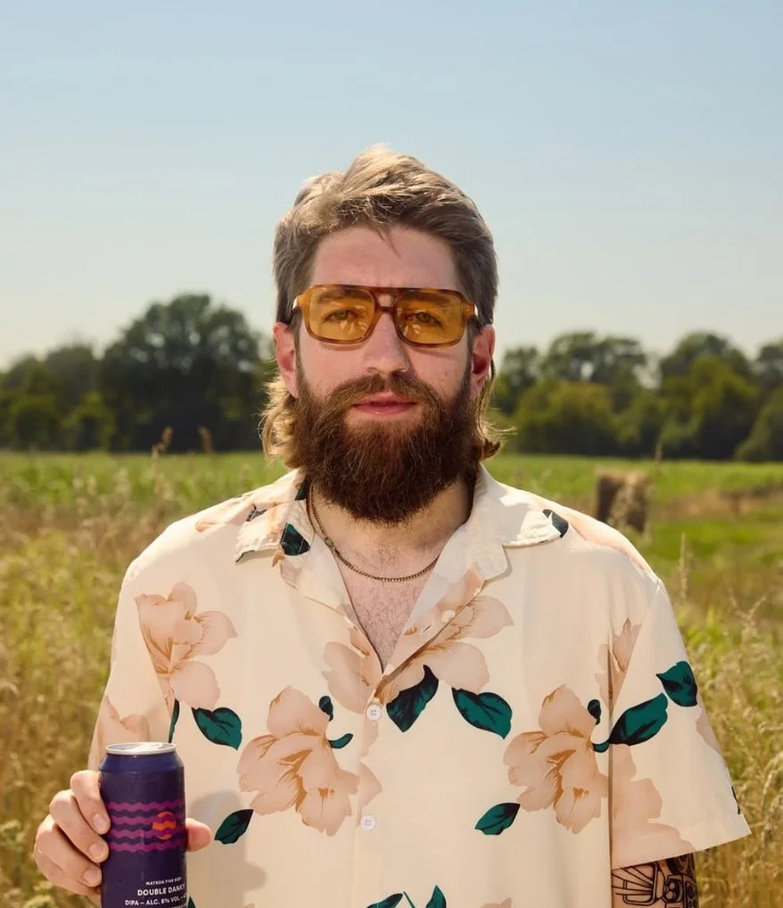 A man with a beard and sunglasses wearing a floral shirt, holding a can, standing outdoors in a grassy field with trees in the background on a sunny day.