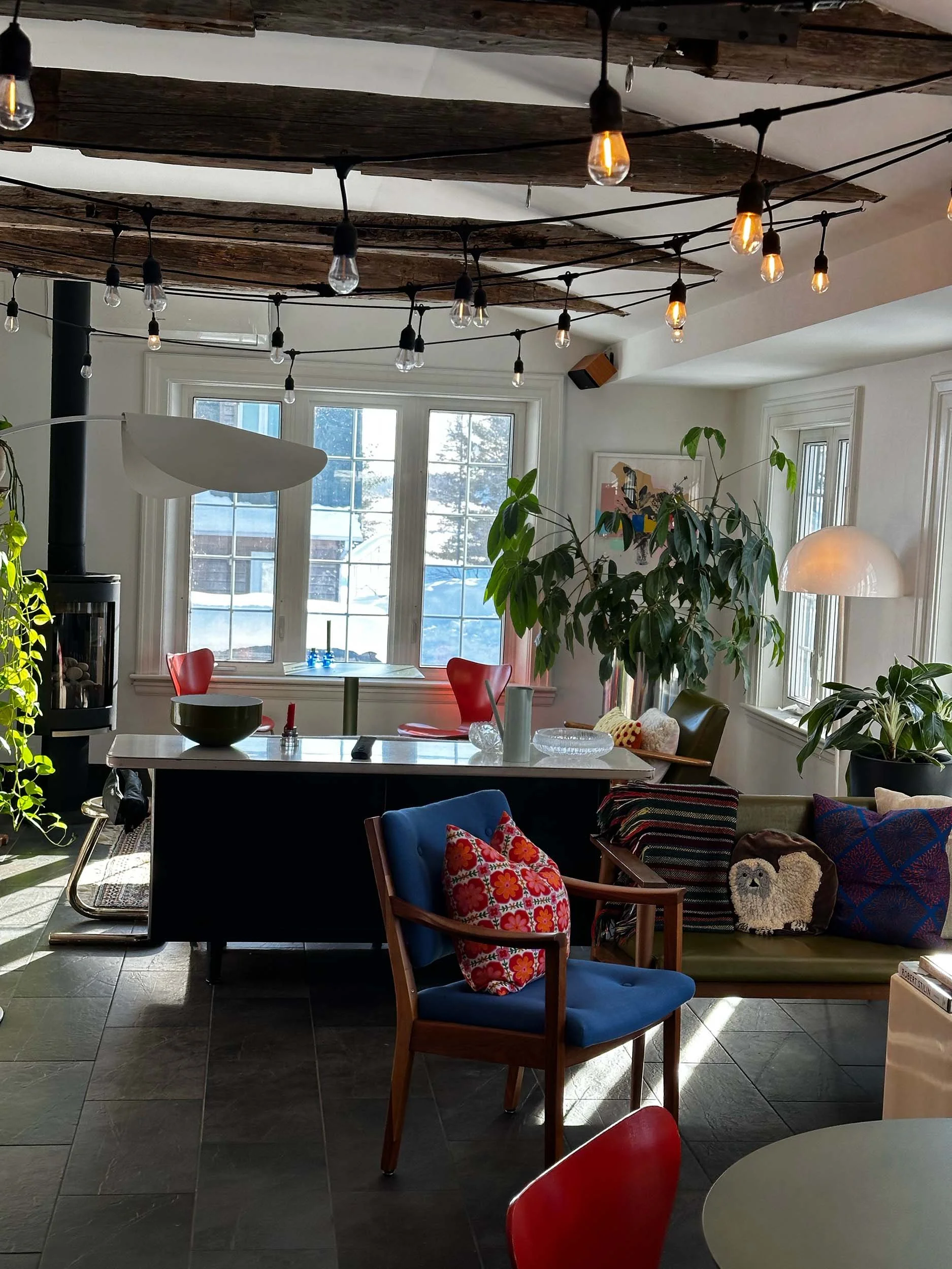 Interior of a cozy cafe space with a mix of modern and vintage furniture, string lights hanging from a wooden ceiling, large window with a view of snowy outdoors, and plants.