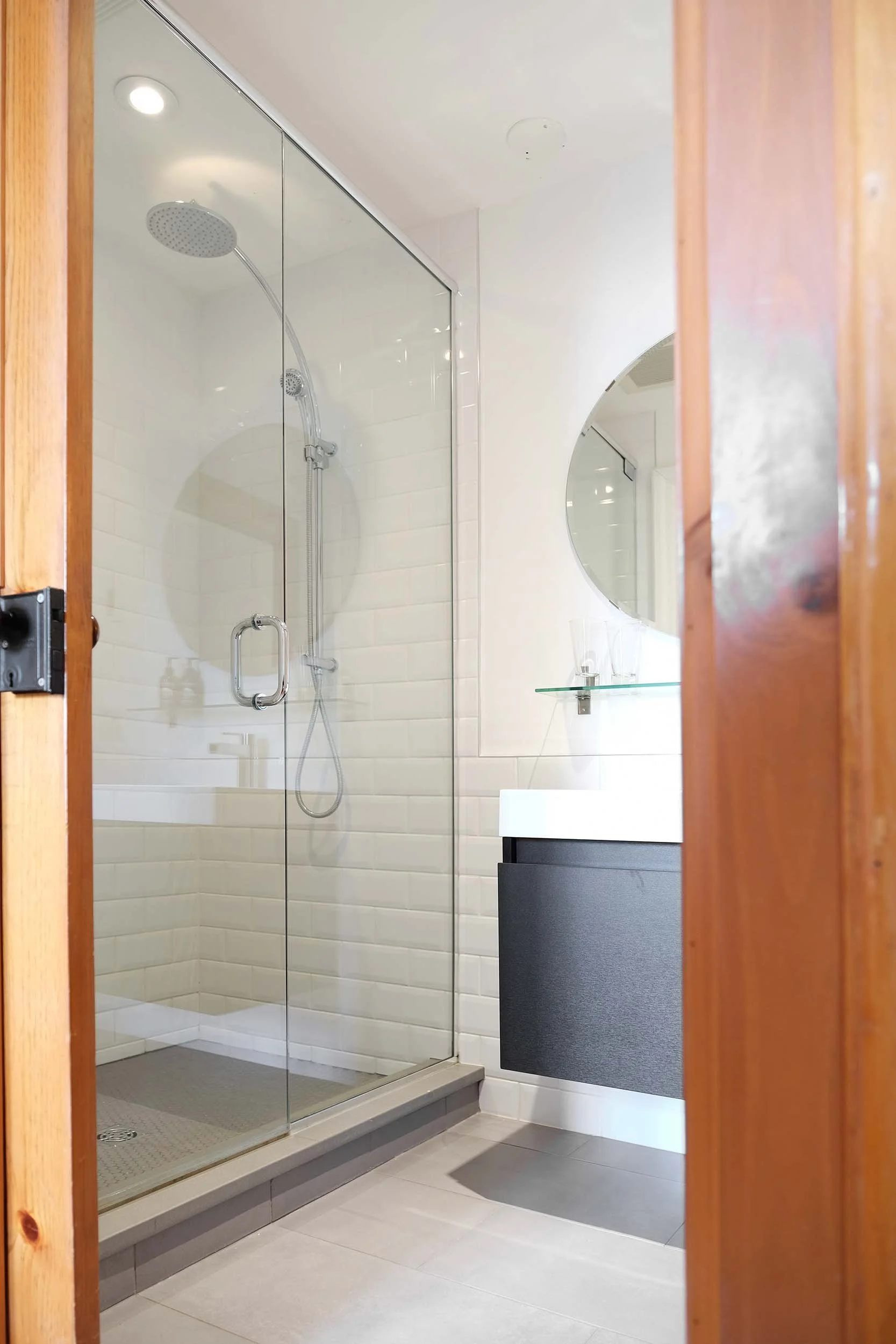 Bathroom with glass shower enclosure, white tiled walls, circular mirror, dark vanity, and wooden door frames.