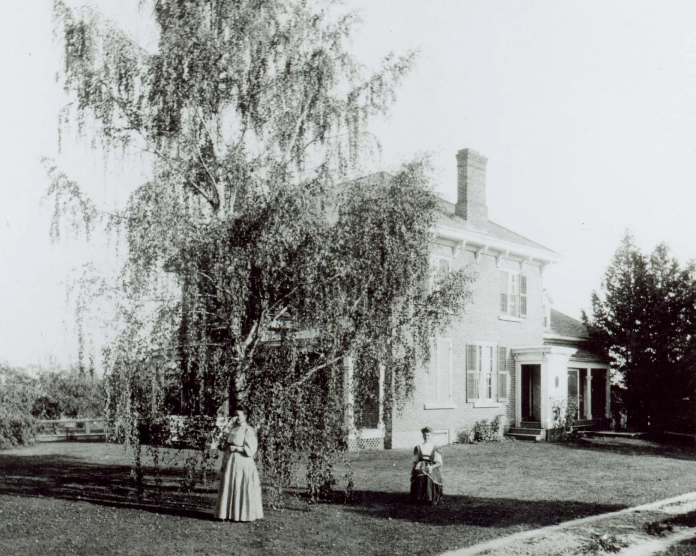 Black and white photo of two women in long dresses standing and sitting in front of a large house with trees nearby.