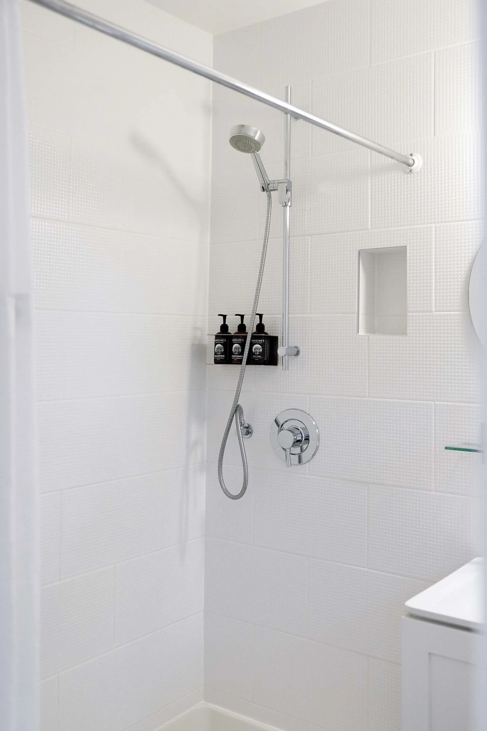 A white tiled shower with a handheld showerhead, a horizontal bar, and three bottles of shampoo, conditioner, and body wash on a wall-mounted shelf.
