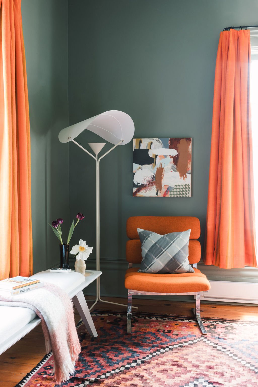 A cozy interior with green walls, orange curtains, an orange chair with a plaid pillow, a white side table with a vase of flowers, a colorful abstract painting, and a unique white floor lamp.