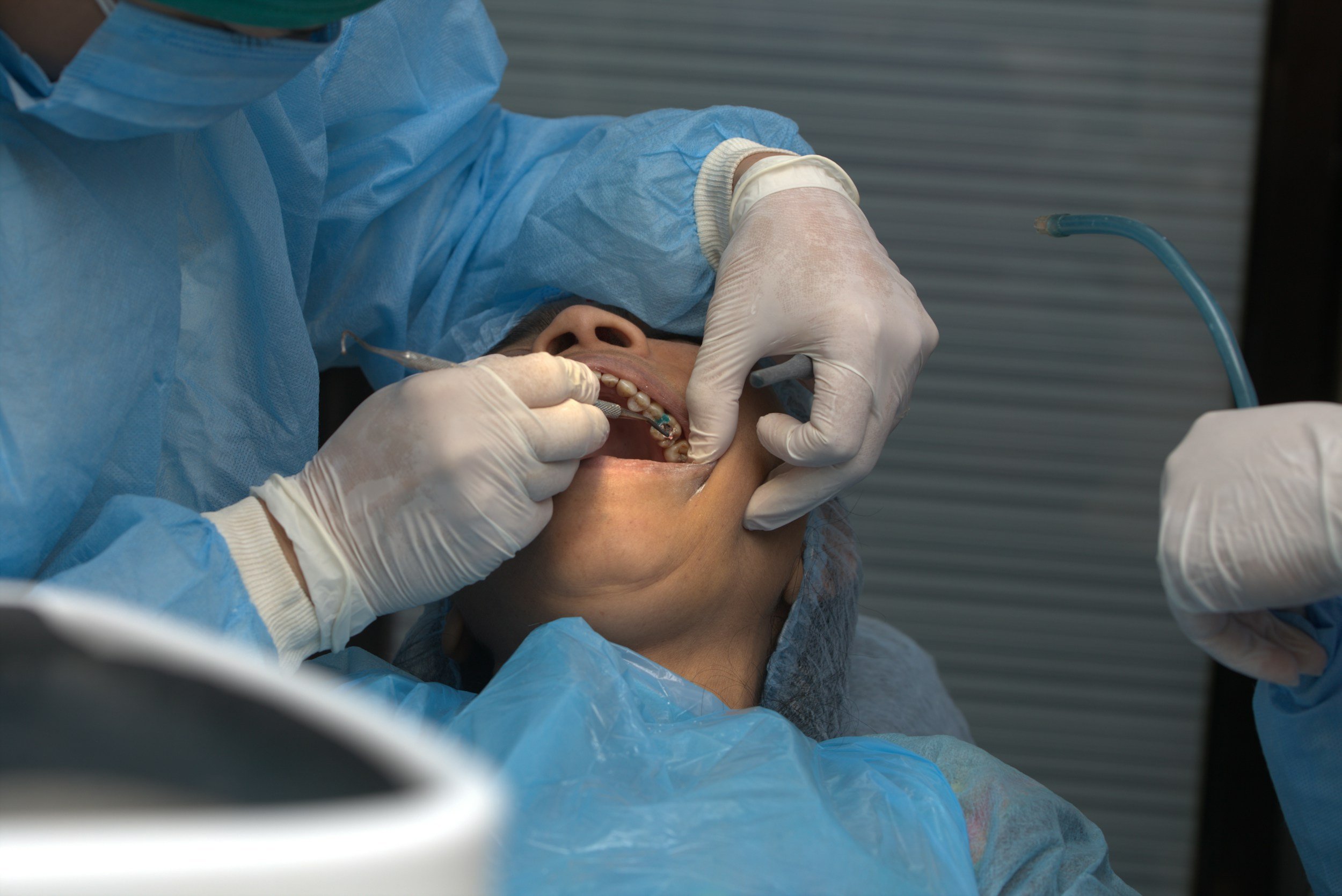 Medical professional in scrubs and gloves performing a dental procedure on a patient lying down, with dental equipment visible.