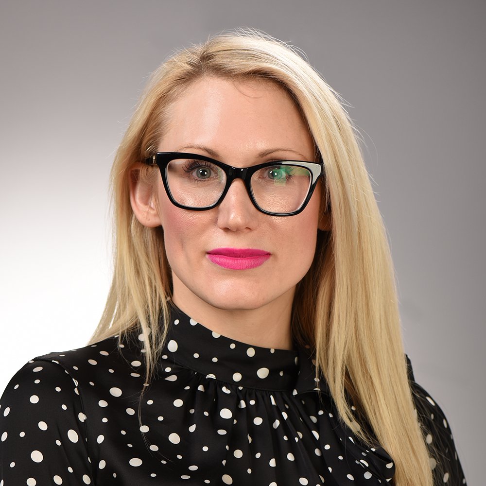 A woman with long blonde hair, wearing glasses, a polka dot black blouse, and pink lipstick, posing against a gray background.