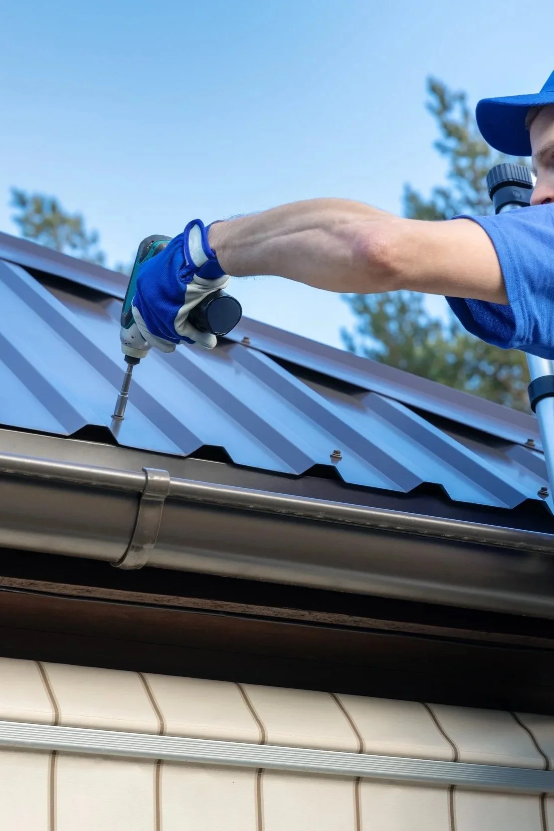 A person wearing a blue shirt, blue gloves, and a blue cap is using a cordless drill to work on a metal roof during daytime with clear blue sky and trees in the background.