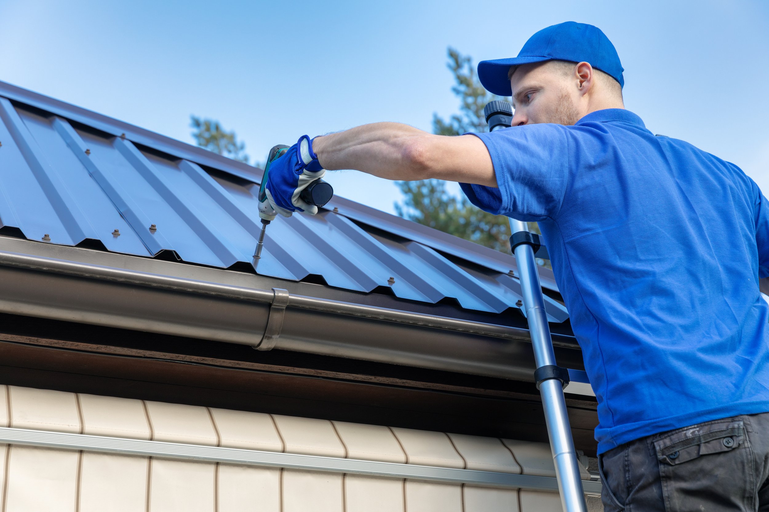 Man wearing blue shirt and cap fixing metal roof shingles with a tool on house.