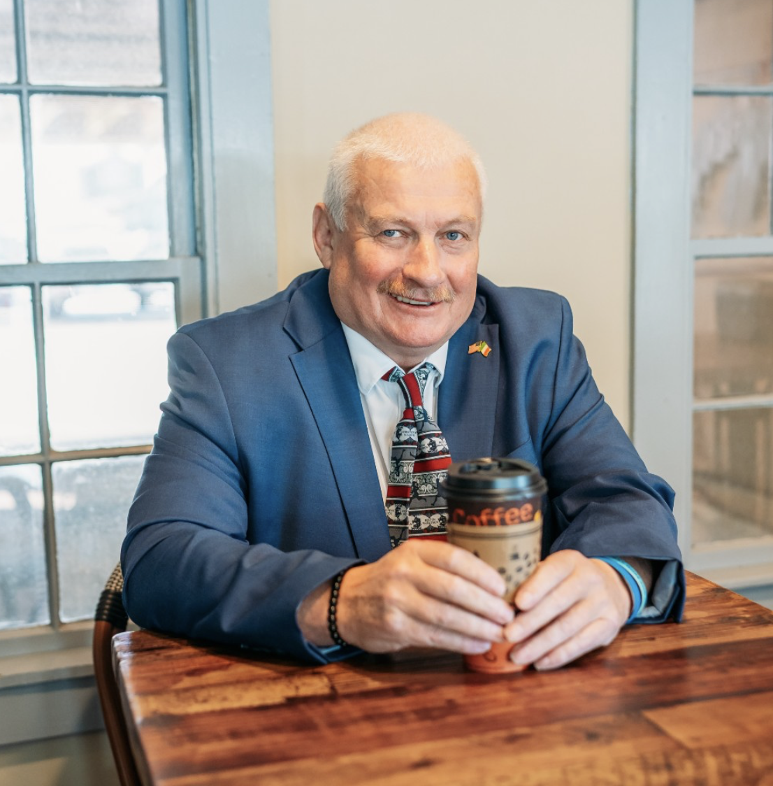 Gerry Connolly sitting at a wooden table, holding a takeout coffee cup with both hands, smiling at the camera. Dressed in a blue suit, white shirt, and tie, with a small flag pin on his lapel, in a bright room with a window.