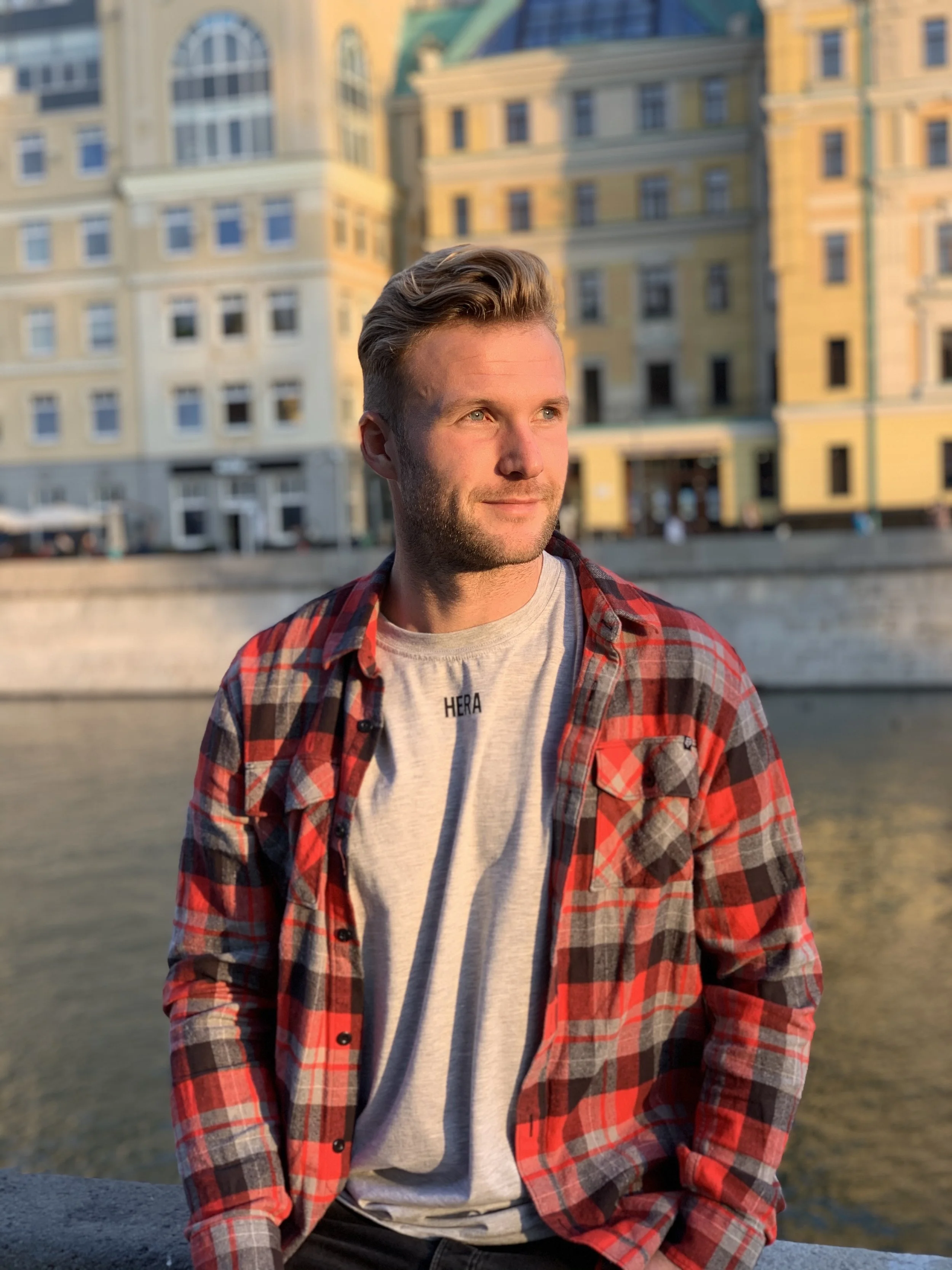 Jongen met blond haar en lichte huid, zittend bij een rivier, met achthoekige gebouwen op de achtergrond tijdens zonsondergang.