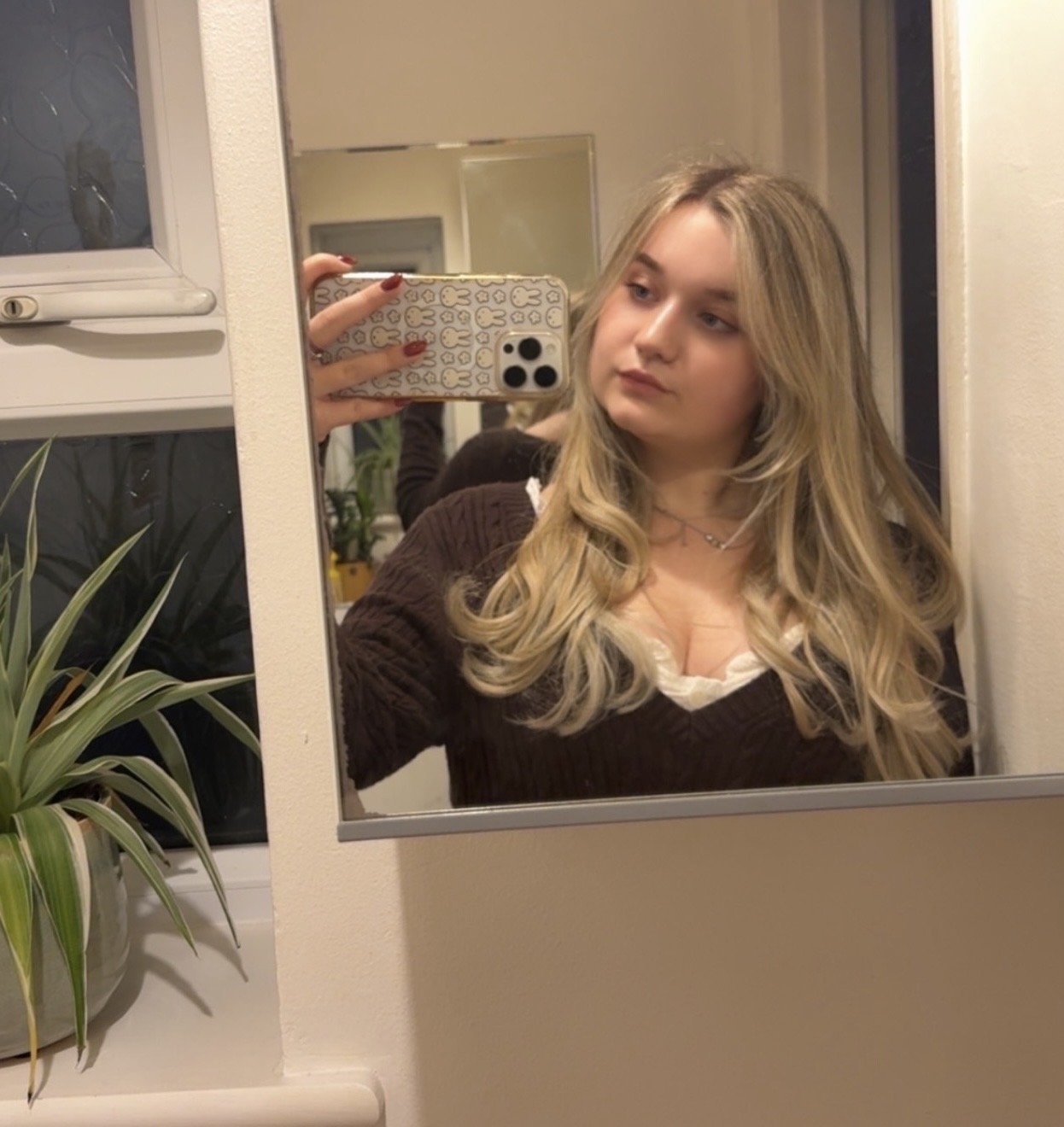 A young woman with long wavy blonde hair taking a mirror selfie in a bathroom, holding a phone with a patterned case featuring cartoon bunnies.