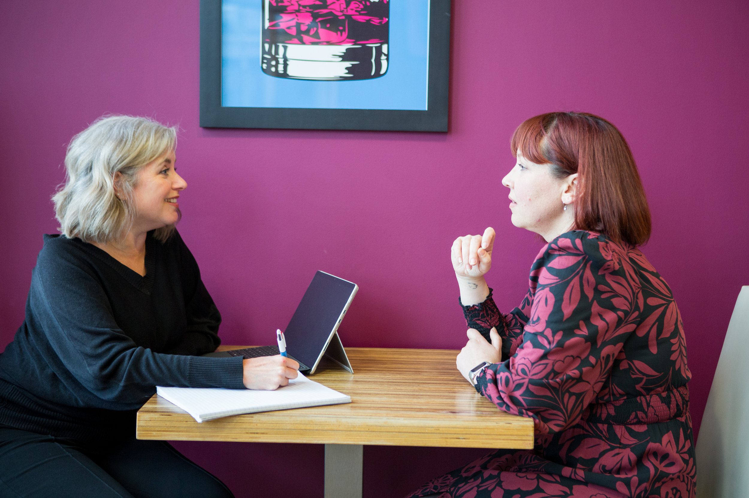 Two women engaging in a conversation at a table in an office or cafe setting, with a purple wall and artwork in the background.