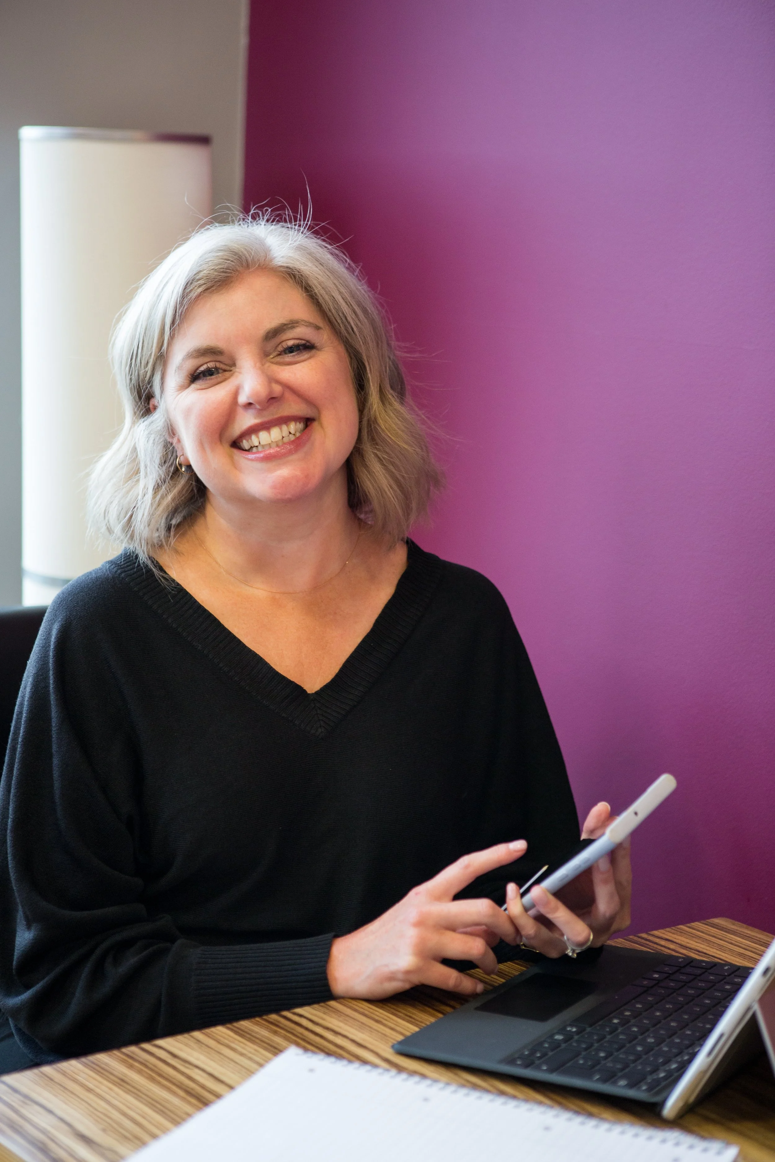 A woman with shoulder-length gray hair smiling and holding a tablet while sitting at a desk with a laptop and a notebook in front of her, against a purple wall.