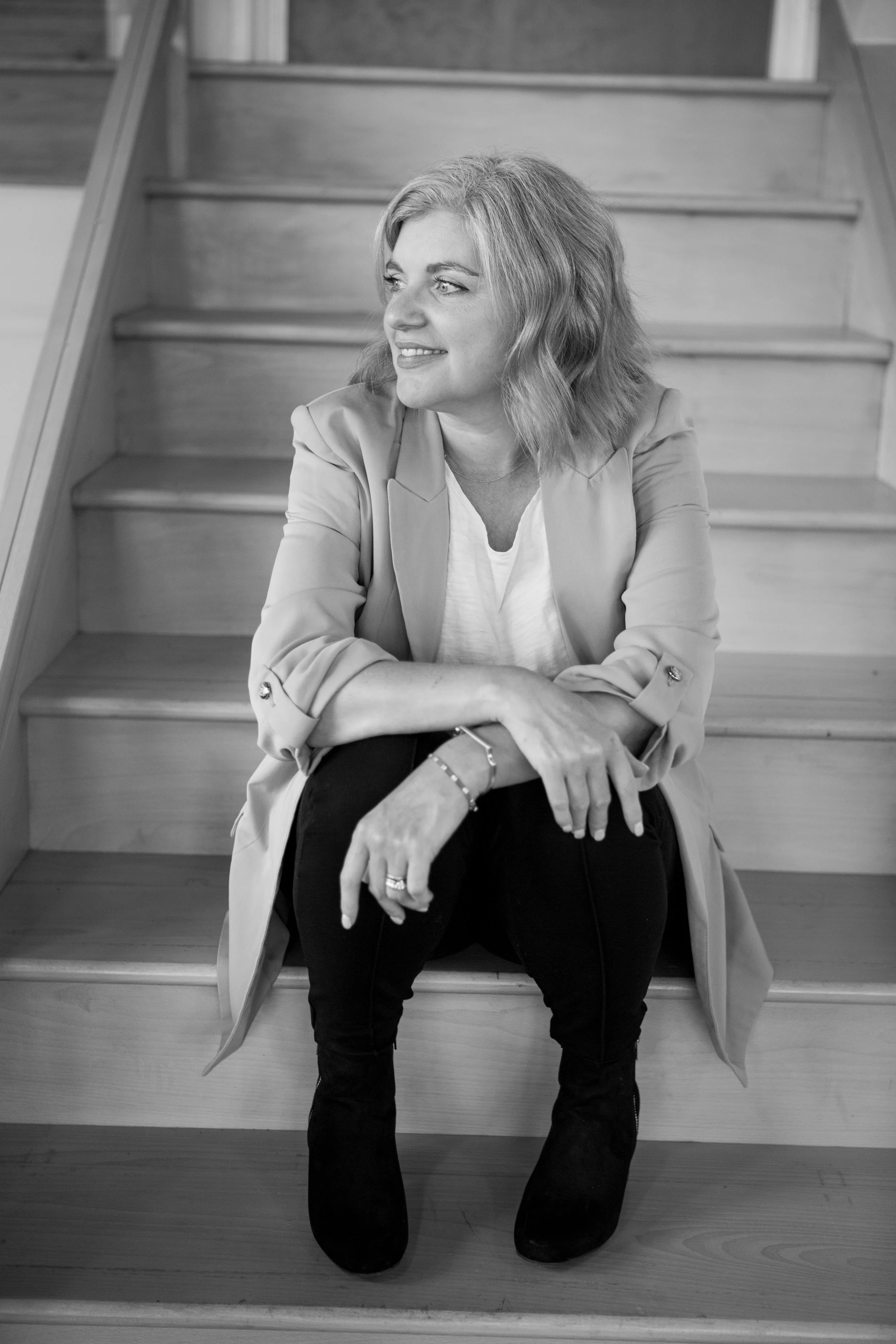 A woman with shoulder-length wavy hair sitting on wooden stairs, smiling and looking to her left. She is wearing a light-colored blazer over a white shirt, black pants, and black boots.