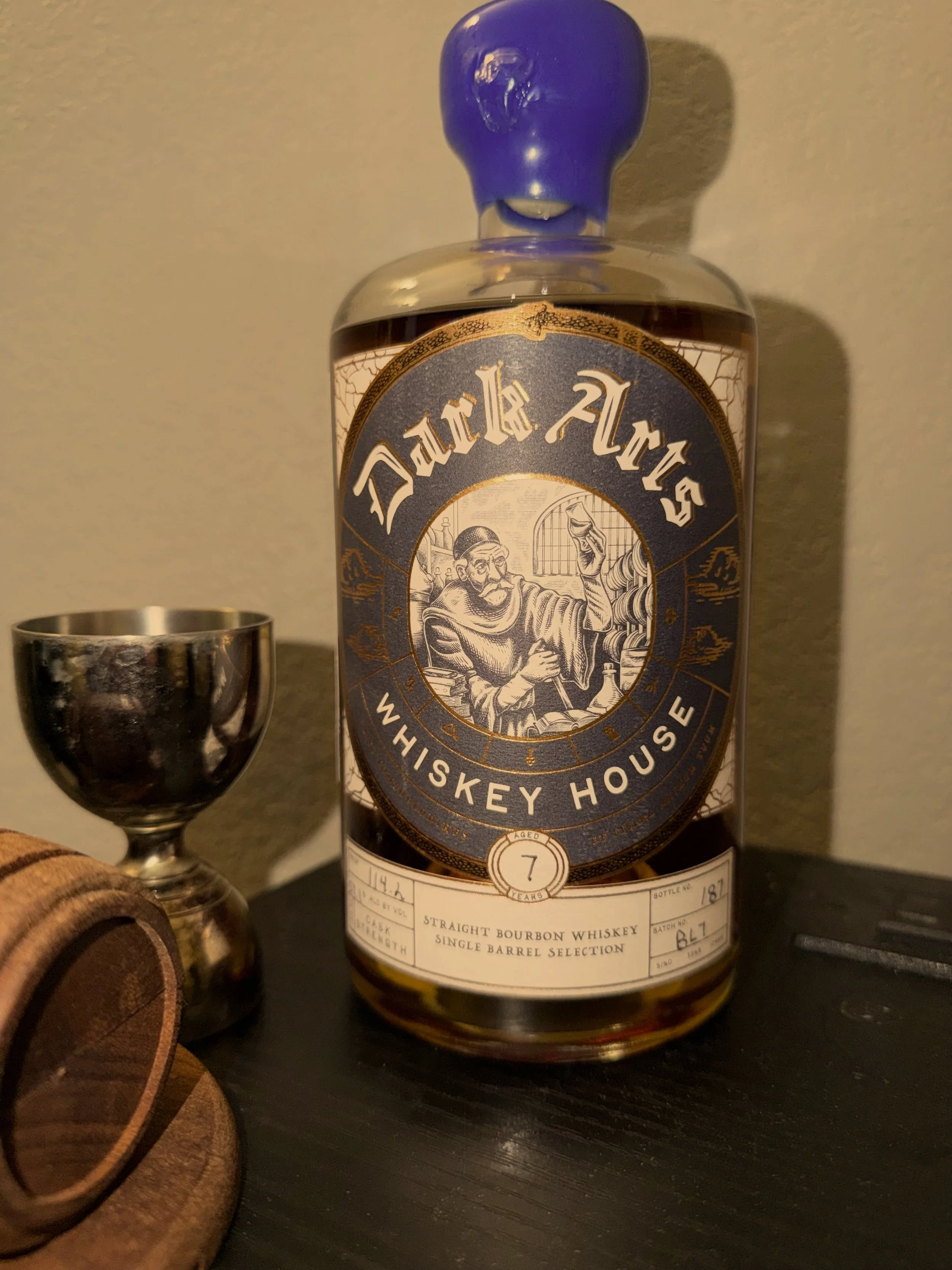 A bottle of Danish Mikes whiskey with a blue cap, alongside a silver shot glass and a small wooden object on a dark surface.