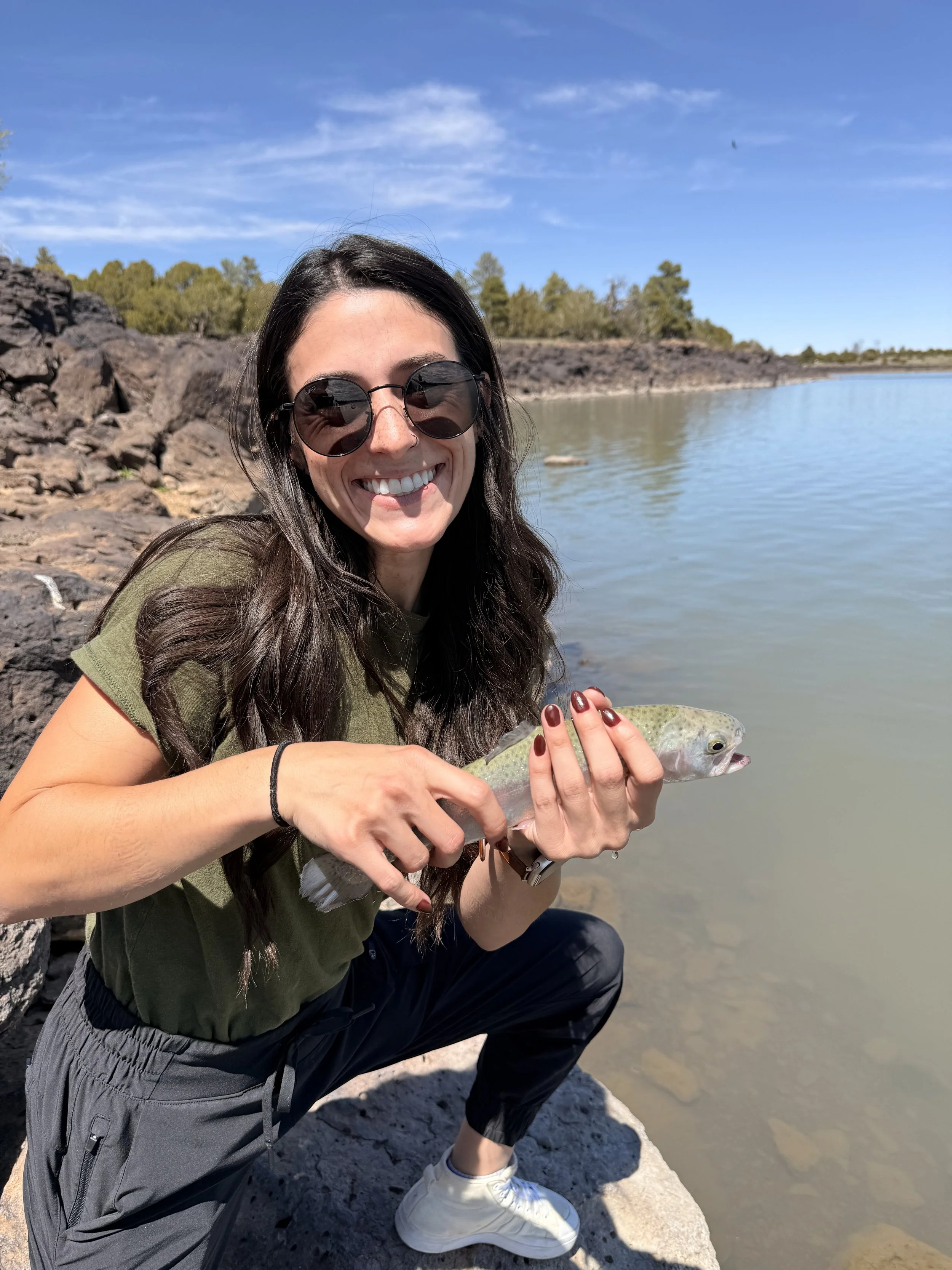 A smiling woman wearing sunglasses kneeling on a rocky shoreline holding a fish she caught near a body of water with trees and a blue sky in the background.