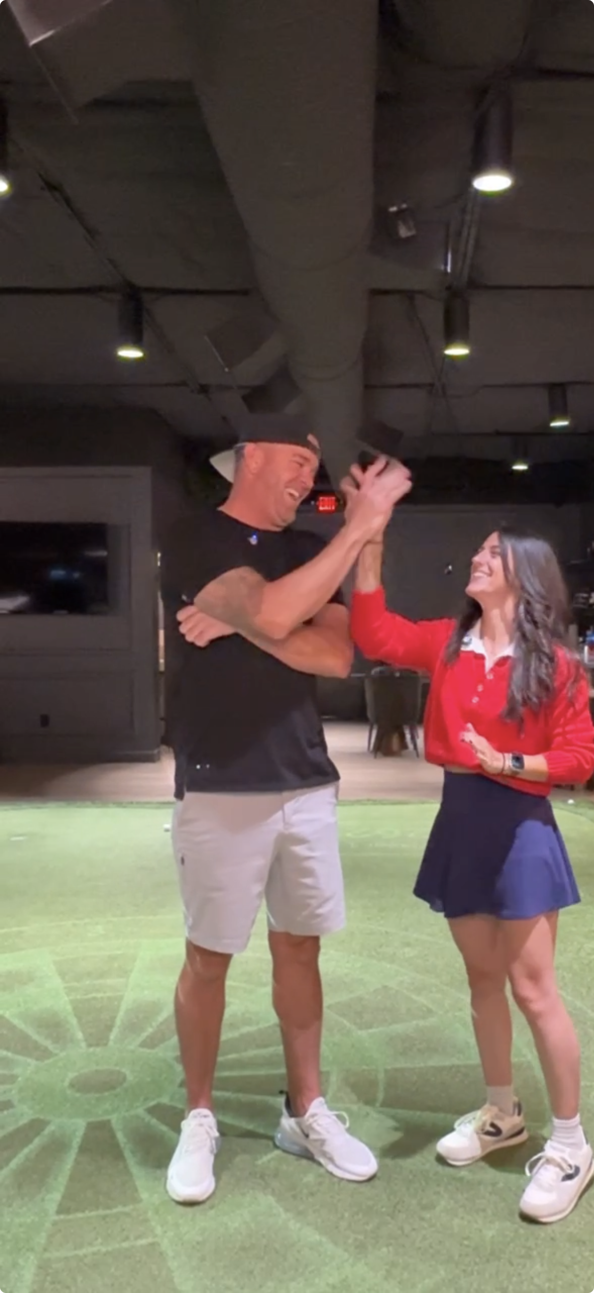 A man and a woman are smiling and laughing, sharing a high five in an indoor setting with green carpeting and ceiling lights.