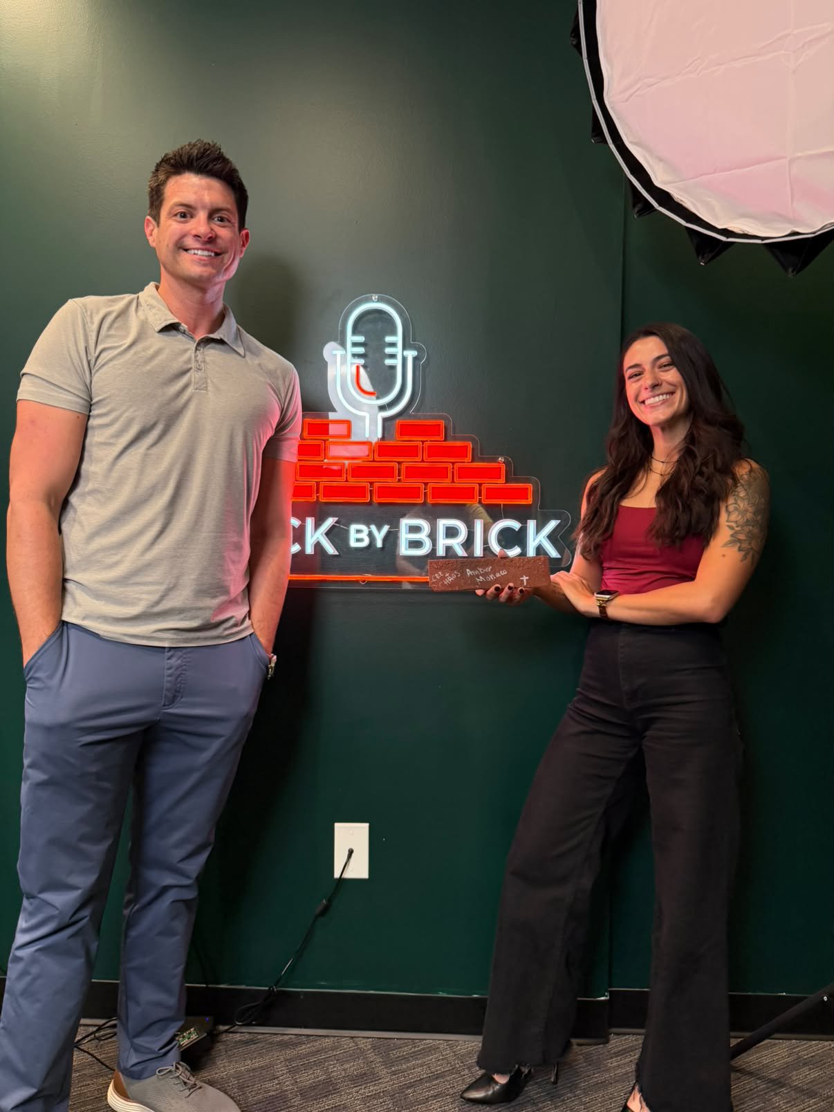 Two people standing in front of a neon sign that reads 'Back by Brick.' The man on the left has short dark hair, a beige polo shirt, and blue pants. The woman on the right has long dark hair, a tattoo on her left arm, and is wearing a burgundy top and black pants, holding a wooden plaque. They are smiling in a room with a dark green wall and professional lighting equipment visible.