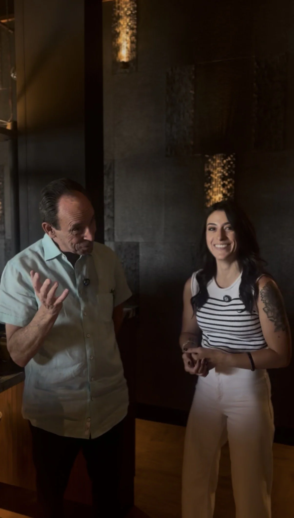 A man and a woman stand indoors, engaged in conversation. The man is gesturing with his hand and wearing a light-colored short-sleeve shirt, while the woman is smiling, wearing a striped sleeveless top and light-colored pants, with dark hair and tattoos on her arm.