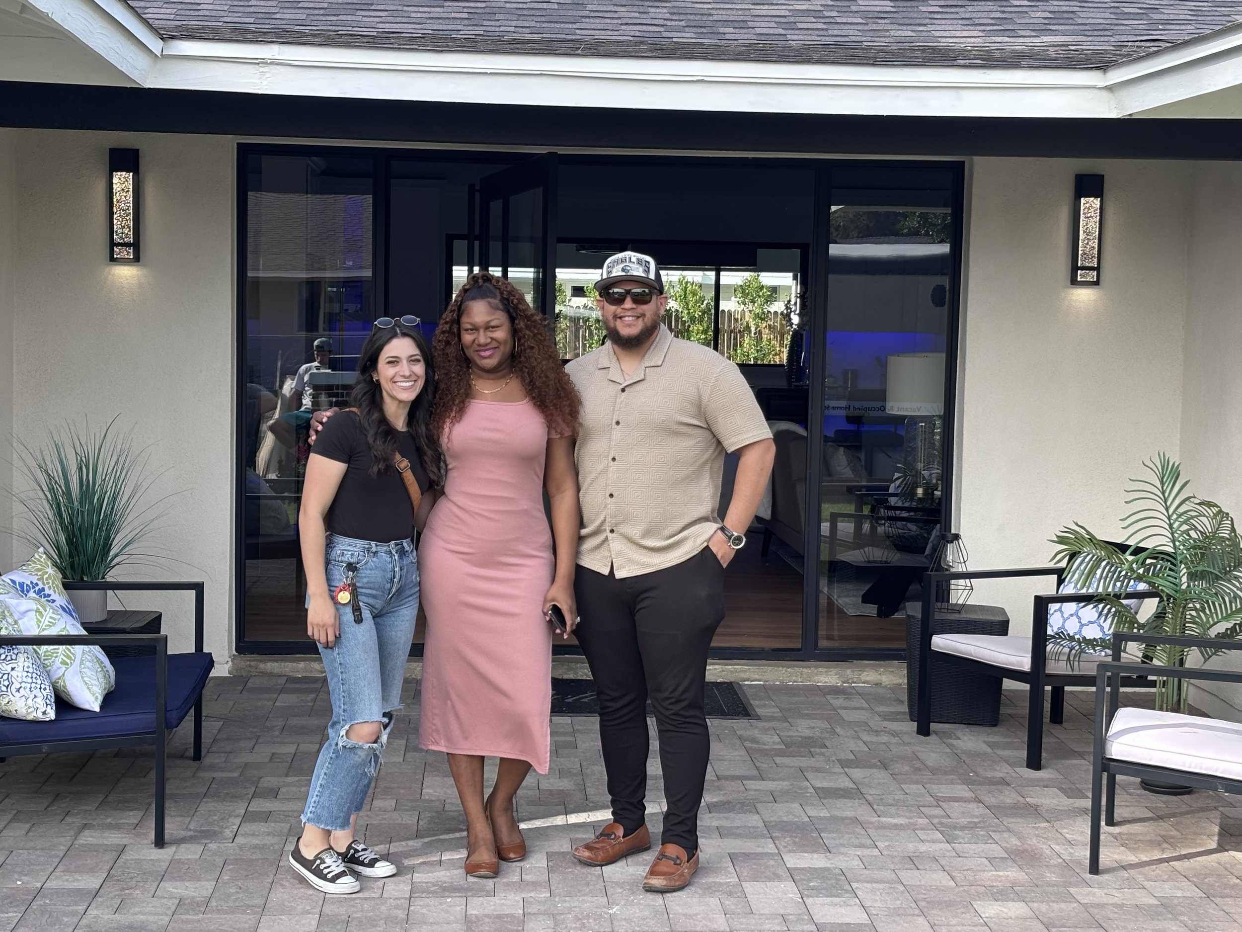 Three people standing together outdoors on a patio in front of a glass door, smiling and posing for a photo. The group consists of one woman in a pink dress and two others, a woman in a black t-shirt and jeans, and a man in a beige shirt and black pants, with trees and a wooden fence visible in the background.
