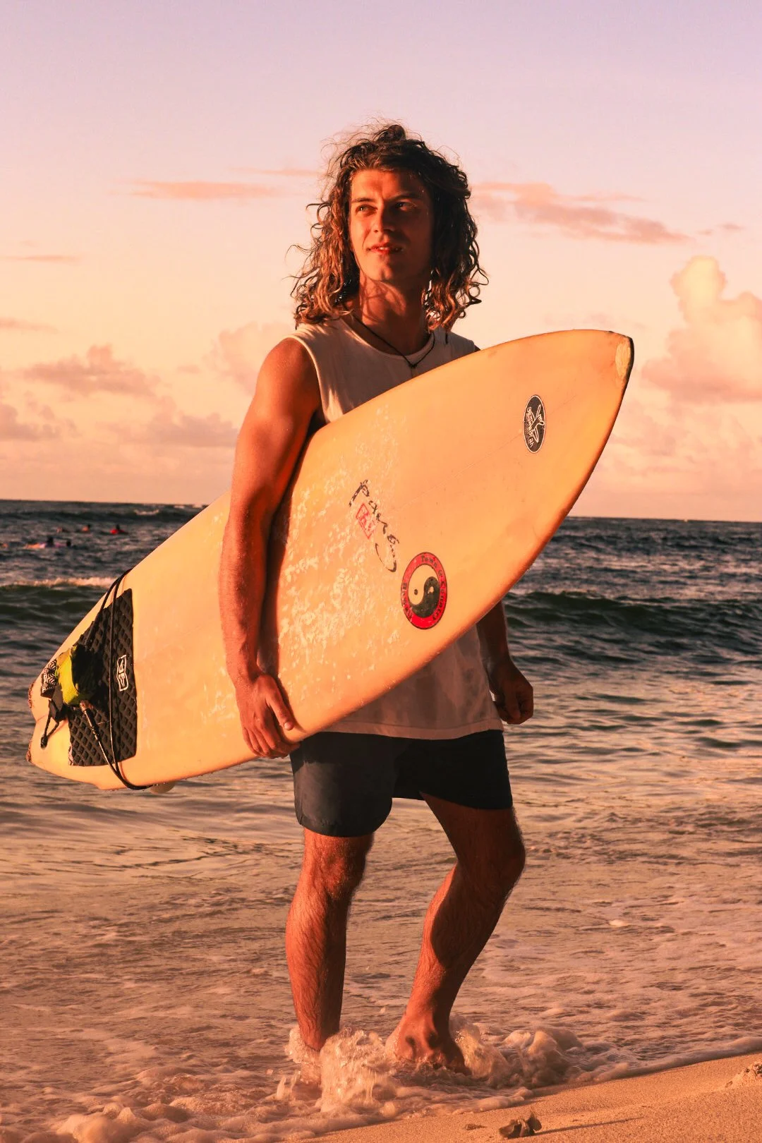 A young man with curly hair holding a yellow surfboard while standing in the ocean at sunset.