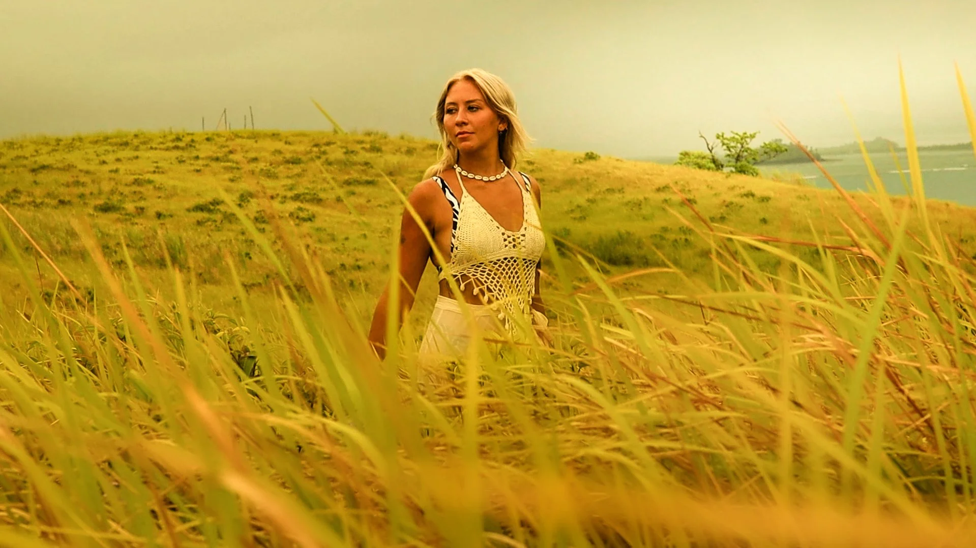 A woman walking through tall grass in a rural landscape with a hill, trees, and a body of water in the background.