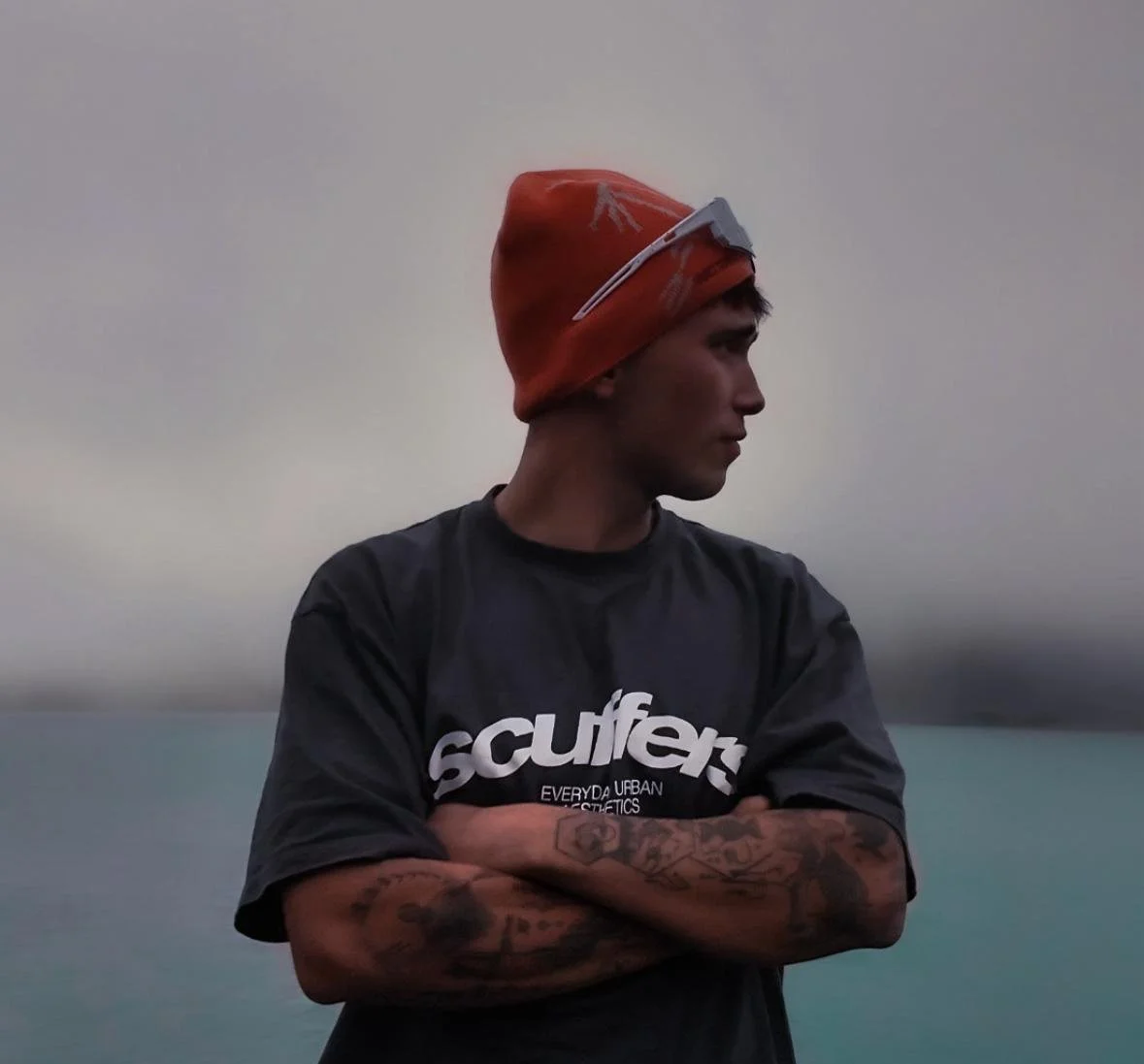 Young man with tattoos on arms wearing a black t-shirt with white text, crossing arms, looking to the side, and wearing an orange beanie with a headlamp on it. Overcast sky in background.