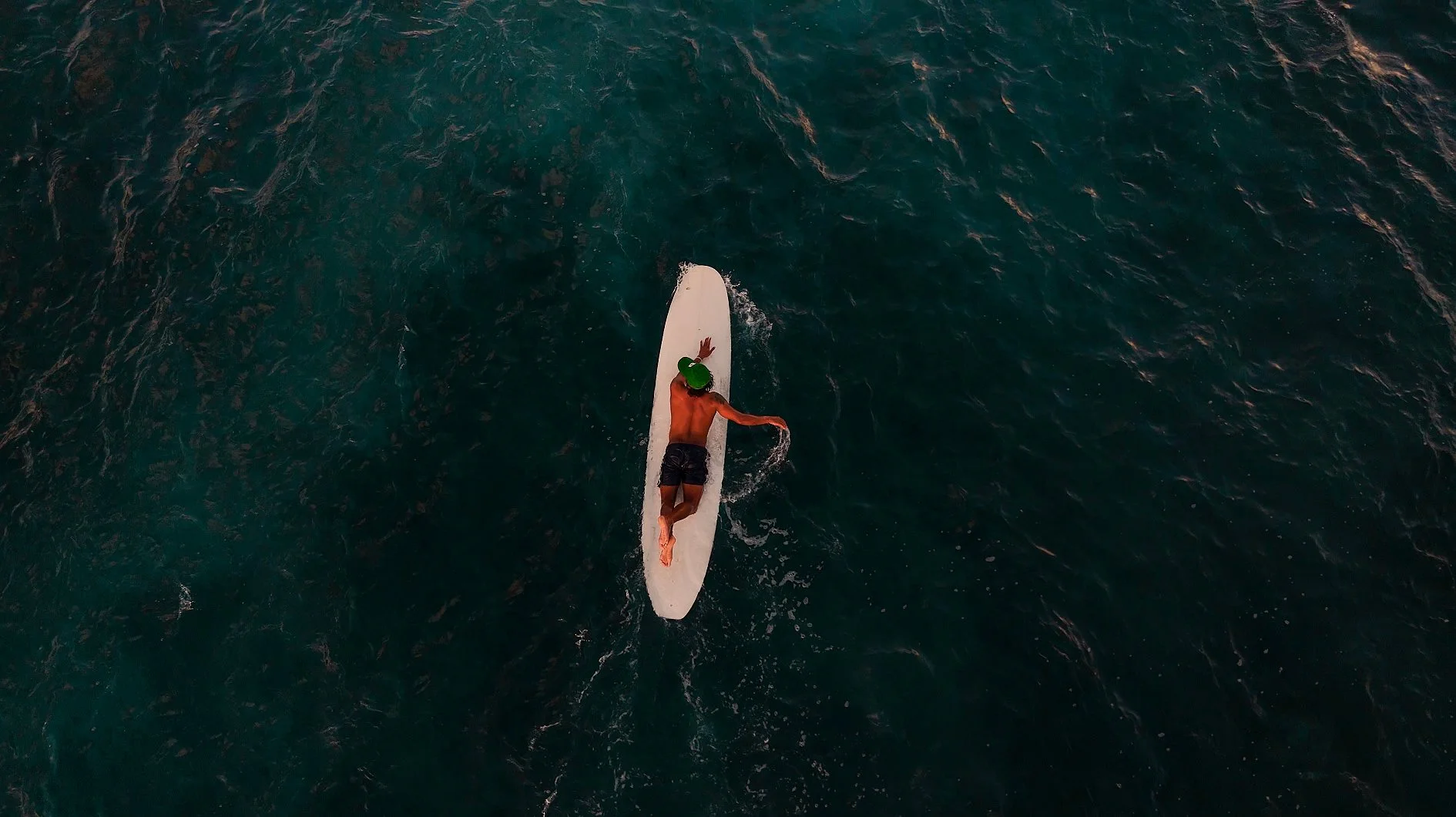 A person surfing on a white surfboard in the ocean, wearing black shorts and a green hat.