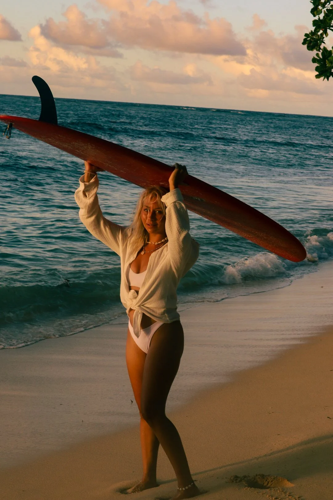 A woman with blonde hair in a bikini and white shirt, holding a surfboard over her head on a beach at sunset.