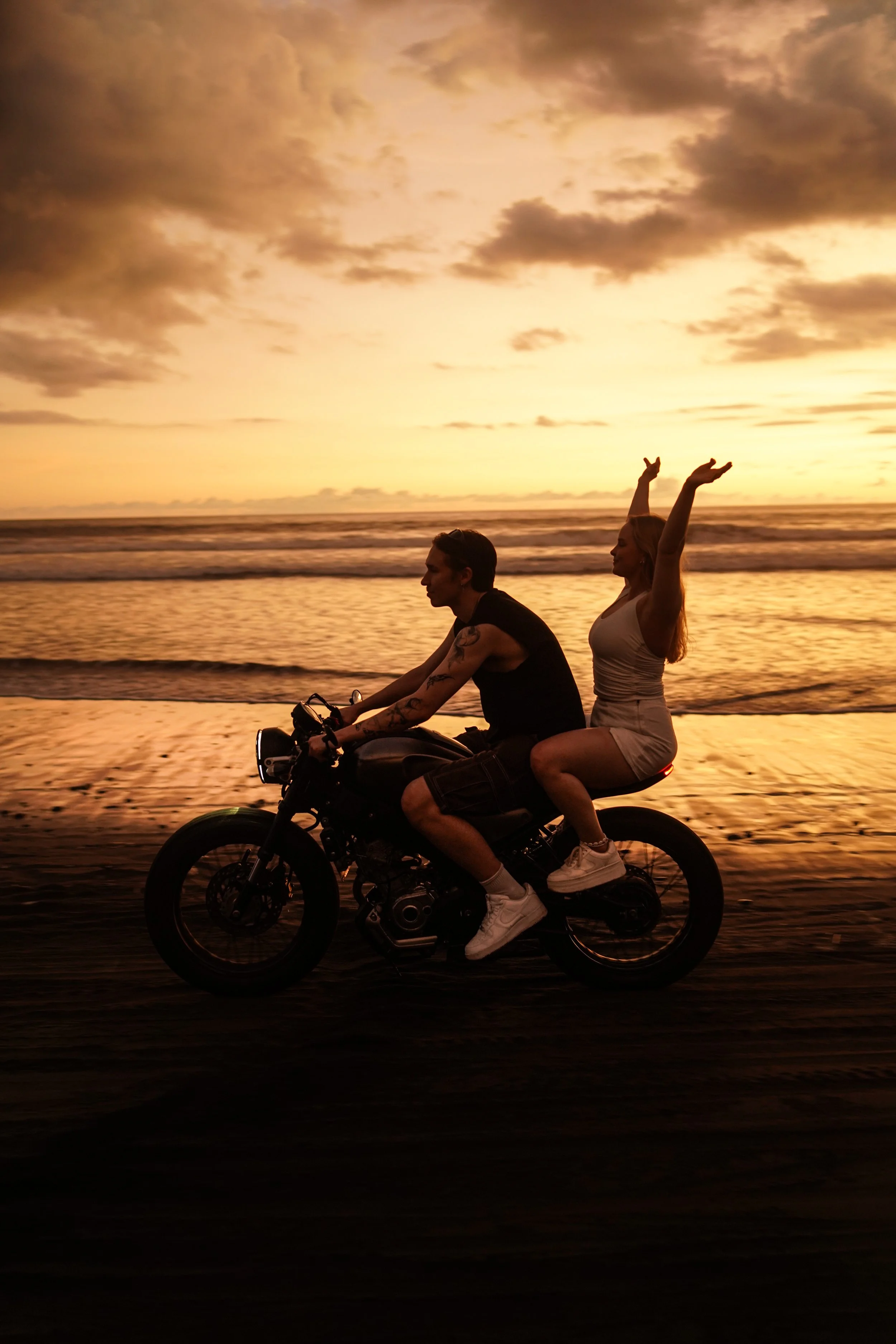 A couple riding a motorcycle along the beach during sunset, with a woman sitting on the back raising her arms in joy.