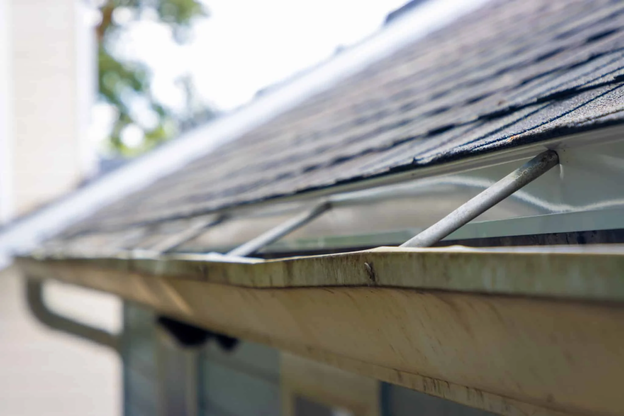 Close-up of a skirting board beneath a window with a metal support rod, house roof with shingles in the background.