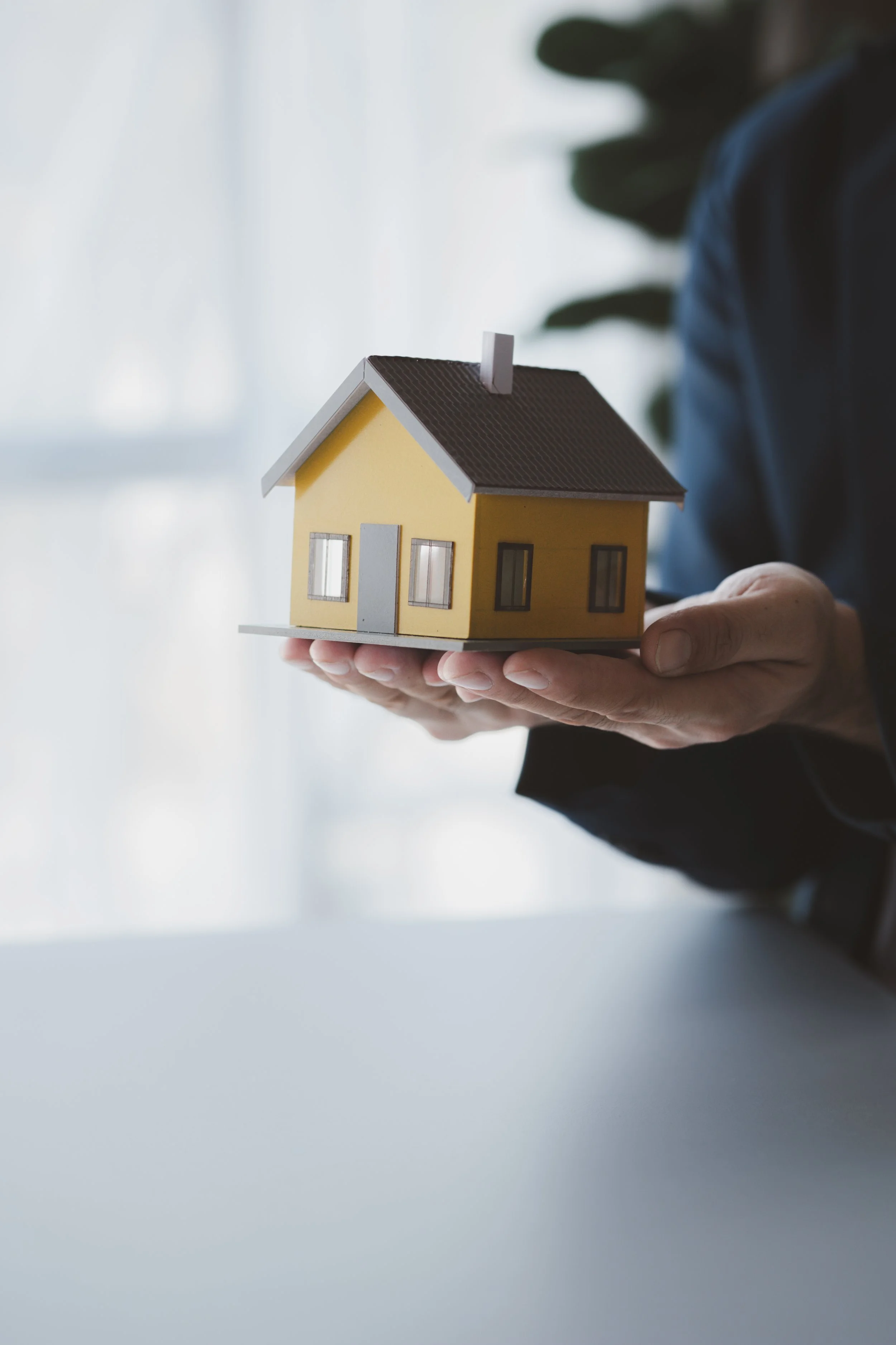 Person holding a small model of a yellow house with gray roof and windows.