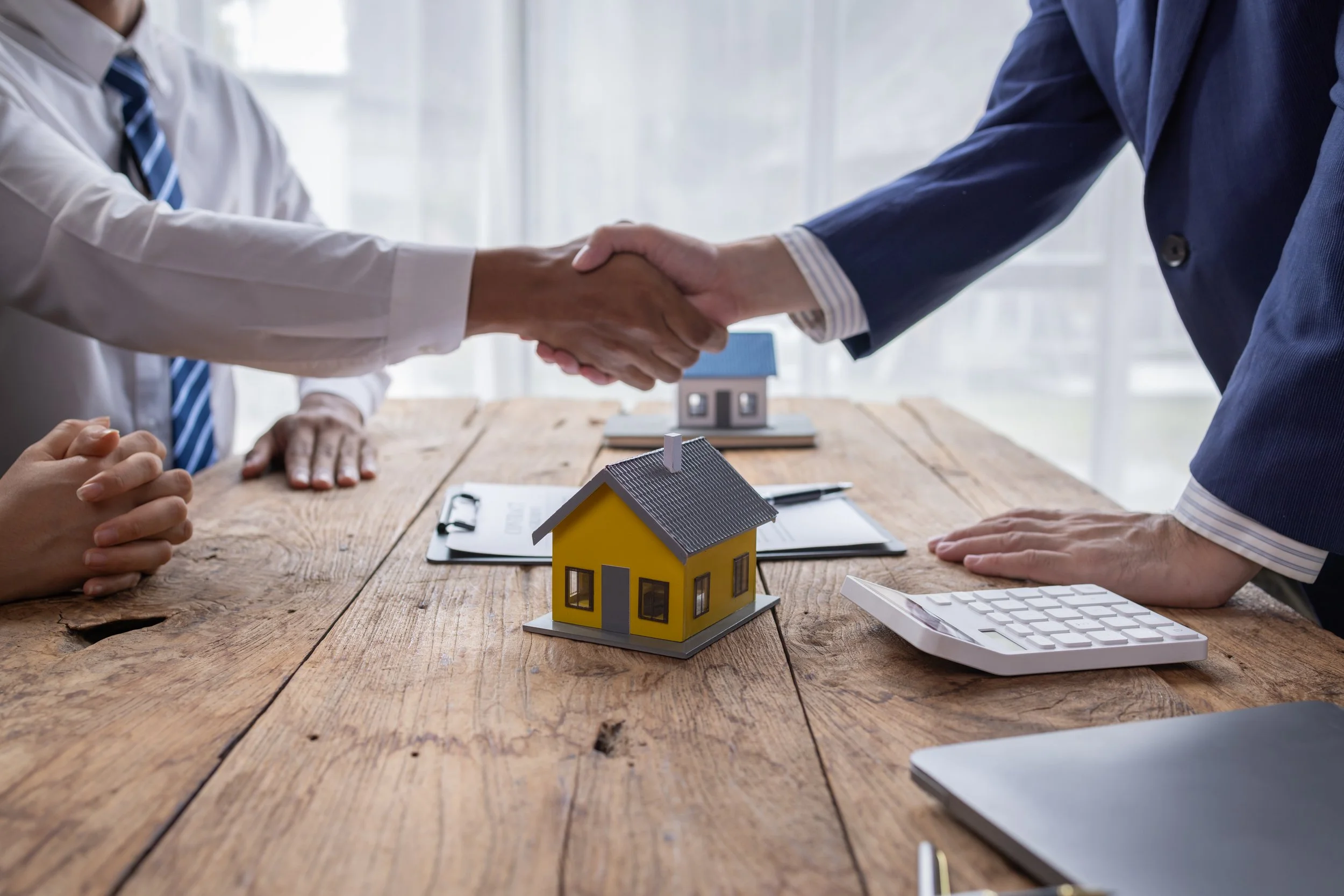 Two people shaking hands over a wooden table with a small yellow house model, documents, a calculator, and another house model in the background, indicating a real estate agreement.