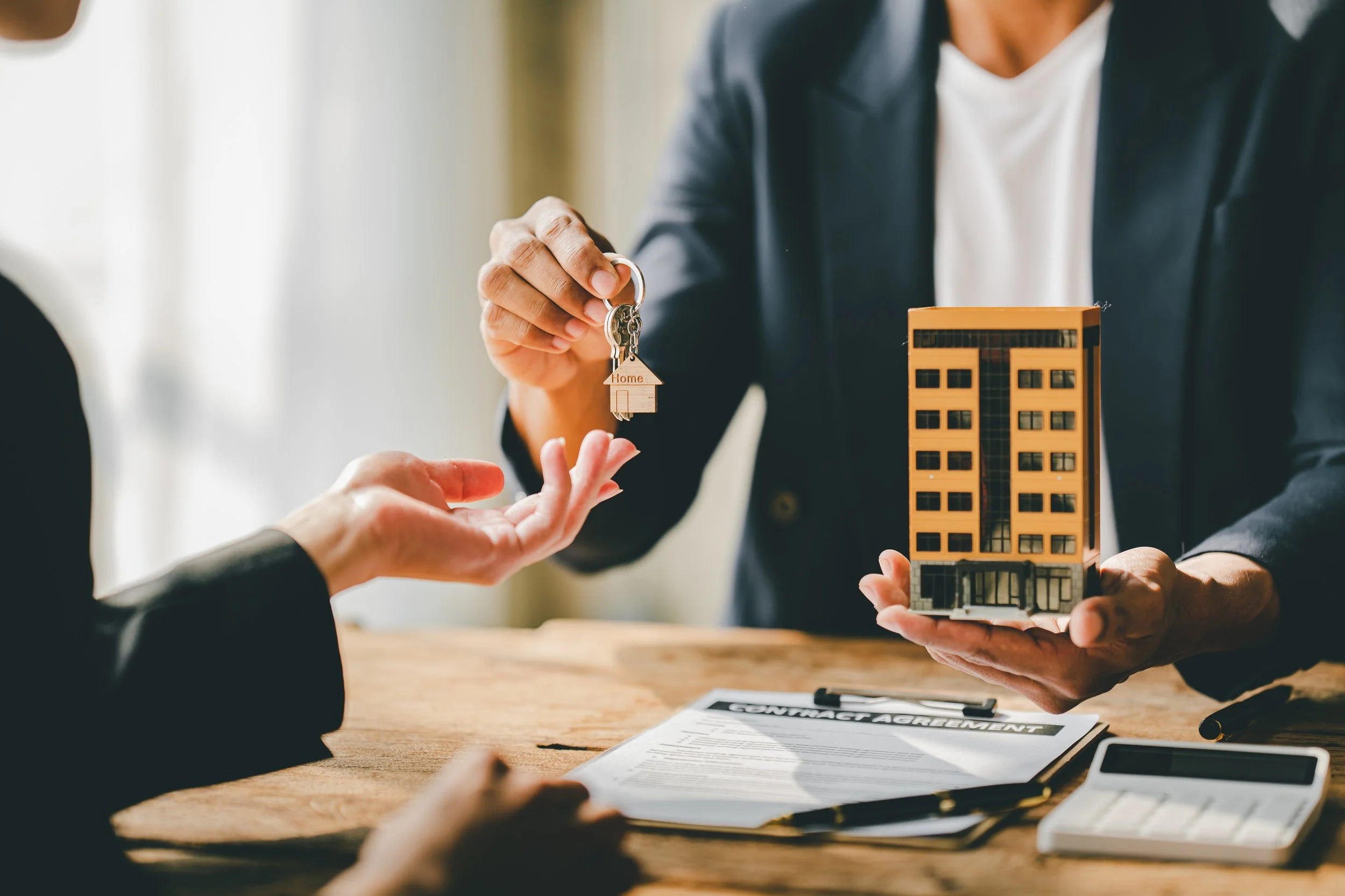 Person handing over house keys to another person at a table with real estate documents, a scale model of a building, and a calculator.