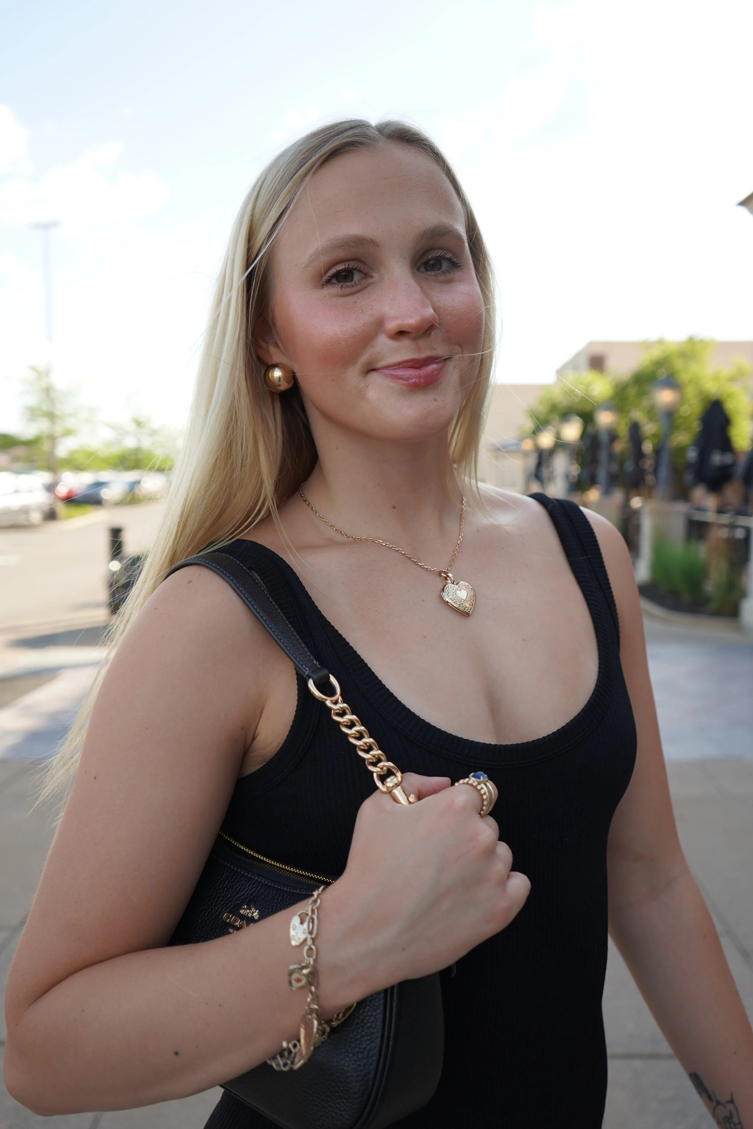 Close-up of a young blonde woman outdoors, wearing a black tank top and gold jewelry, with a cheerful expression and a background of a parking lot and green trees.