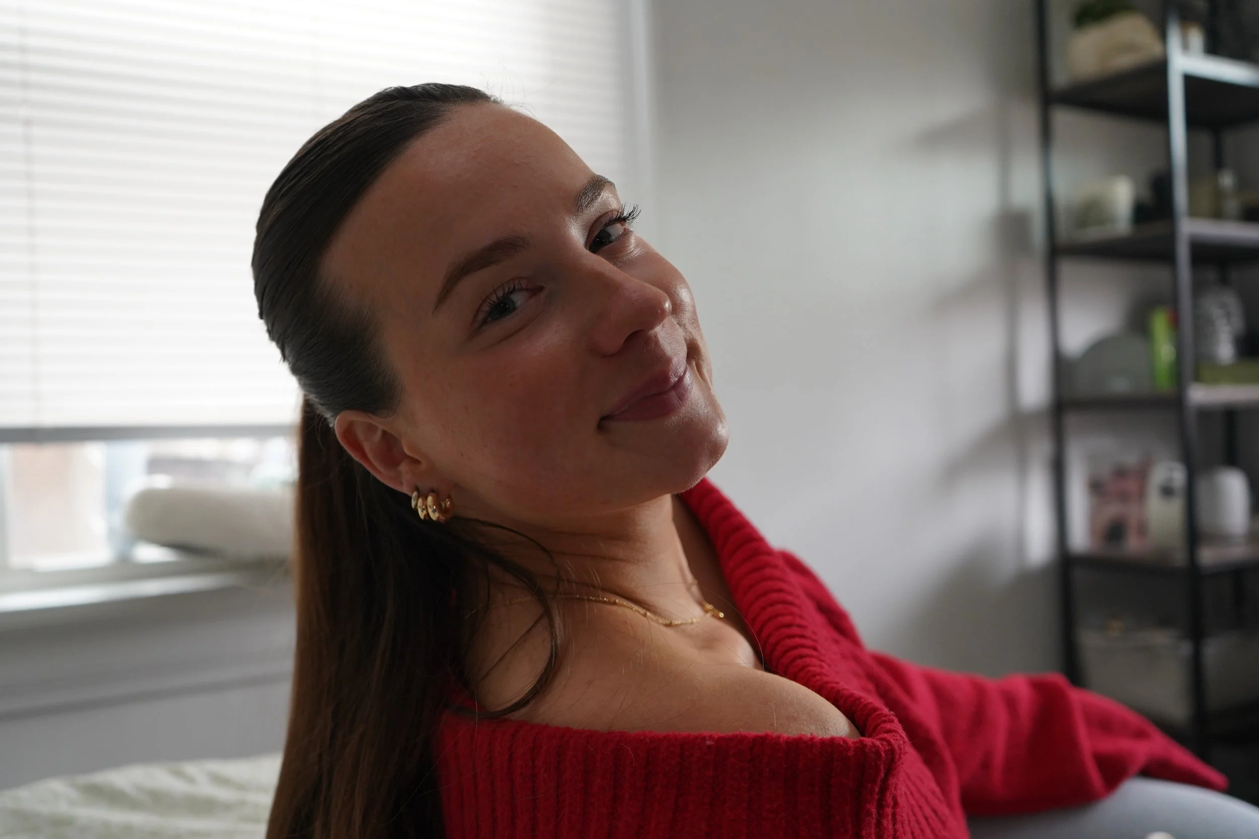 A woman smiling and looking at the camera in a cozy indoor setting with shelves in the background and a window with blinds.