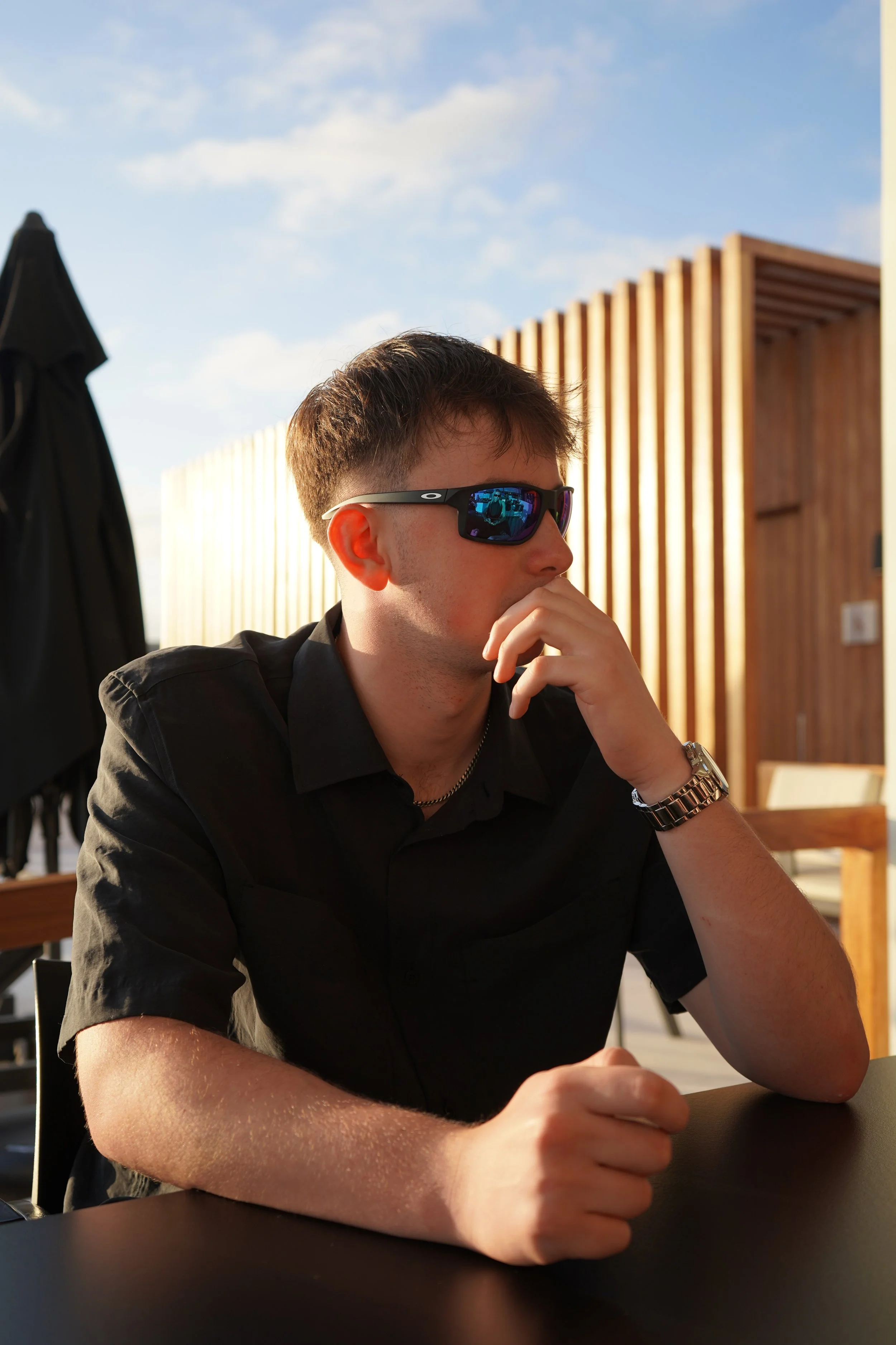 A young man with short brown hair wearing dark sunglasses with a reflective blue mirror tint, a black shirt, a silver watch, and a silver chain, sitting at a table with a thoughtful expression and hand near his face, outdoors with a wooden structure 