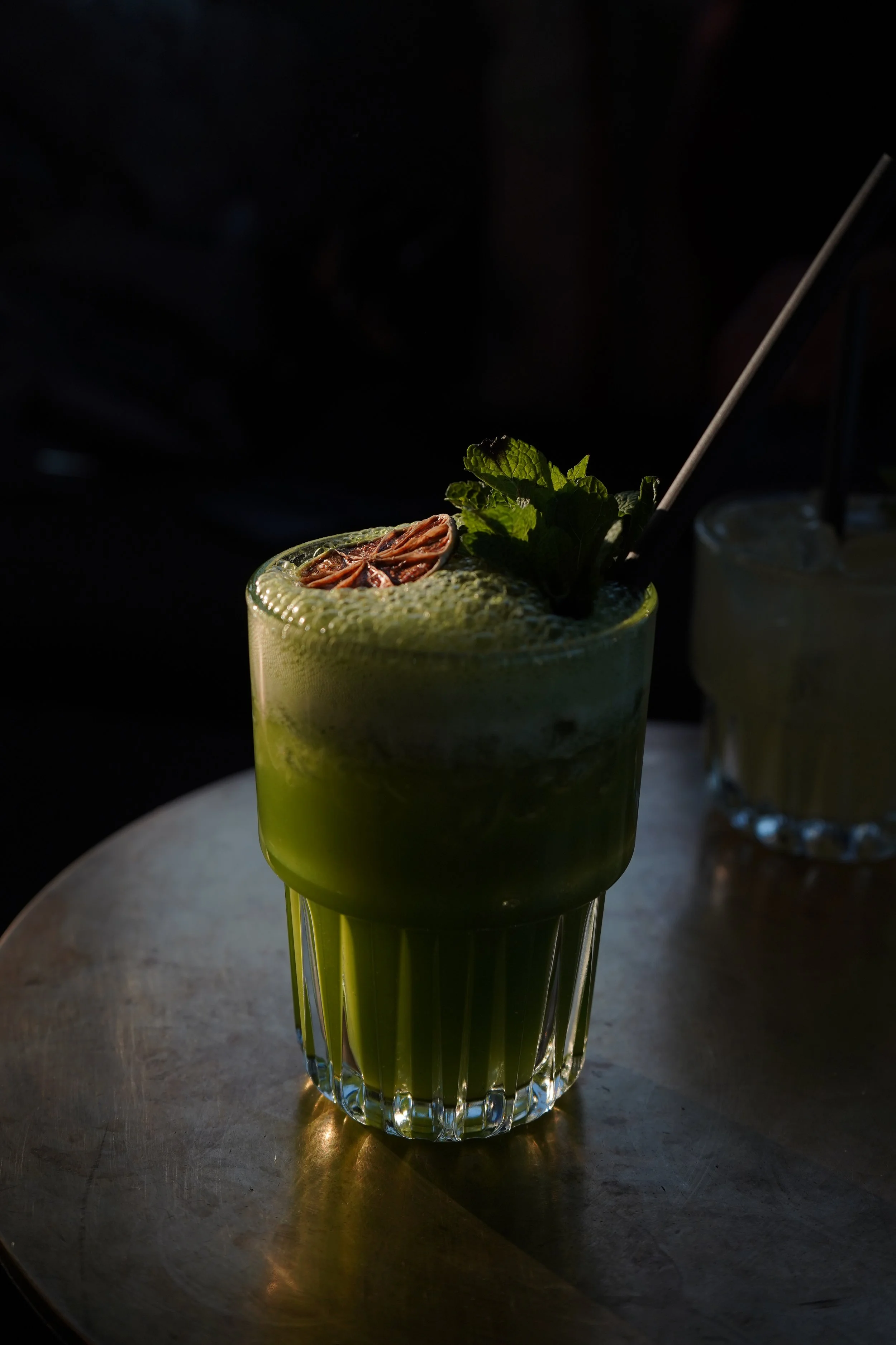 A green matcha cocktail garnished with a dried lime slice and fresh mint leaves, served in a tall clear glass on a metal surface.