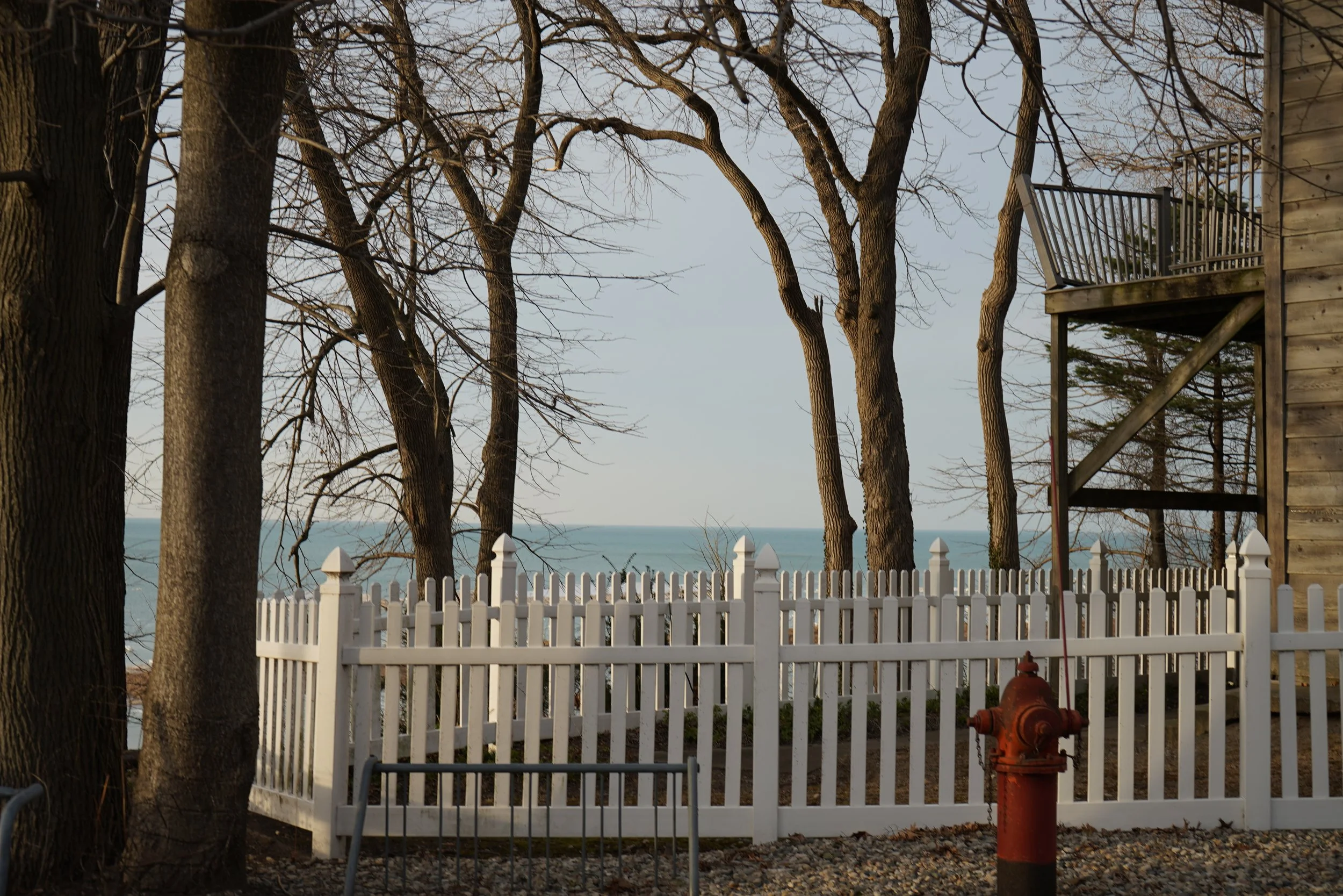Leafless trees behind a white picket fence and a brick building with a balcony, with the ocean visible in the distance.