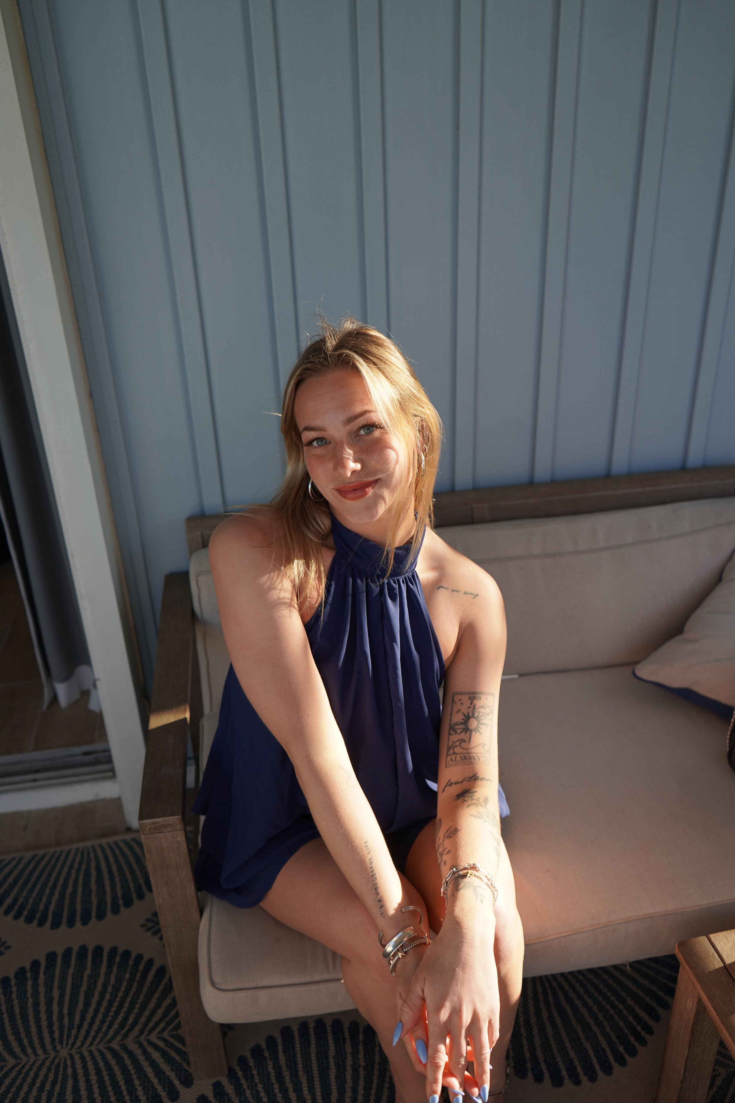 A young woman with blonde hair and tattoos, wearing a sleeveless dark blue dress, sitting on a beige outdoor sofa, smiling at the camera, with a patterned rug and a blue siding wall in the background.
