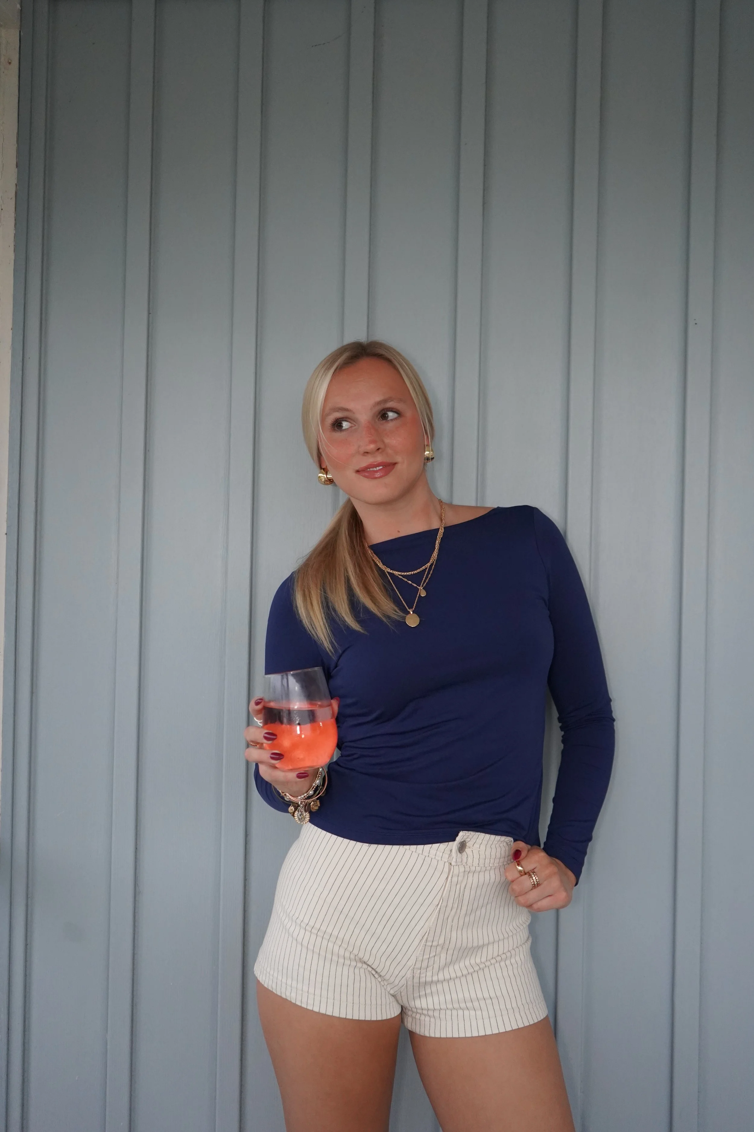 A woman with blonde hair in a ponytail is holding a glass of a pink drink. She is wearing a navy blue long-sleeve top and beige striped shorts, accessorized with gold jewelry. She stands in front of a blue paneled wall.