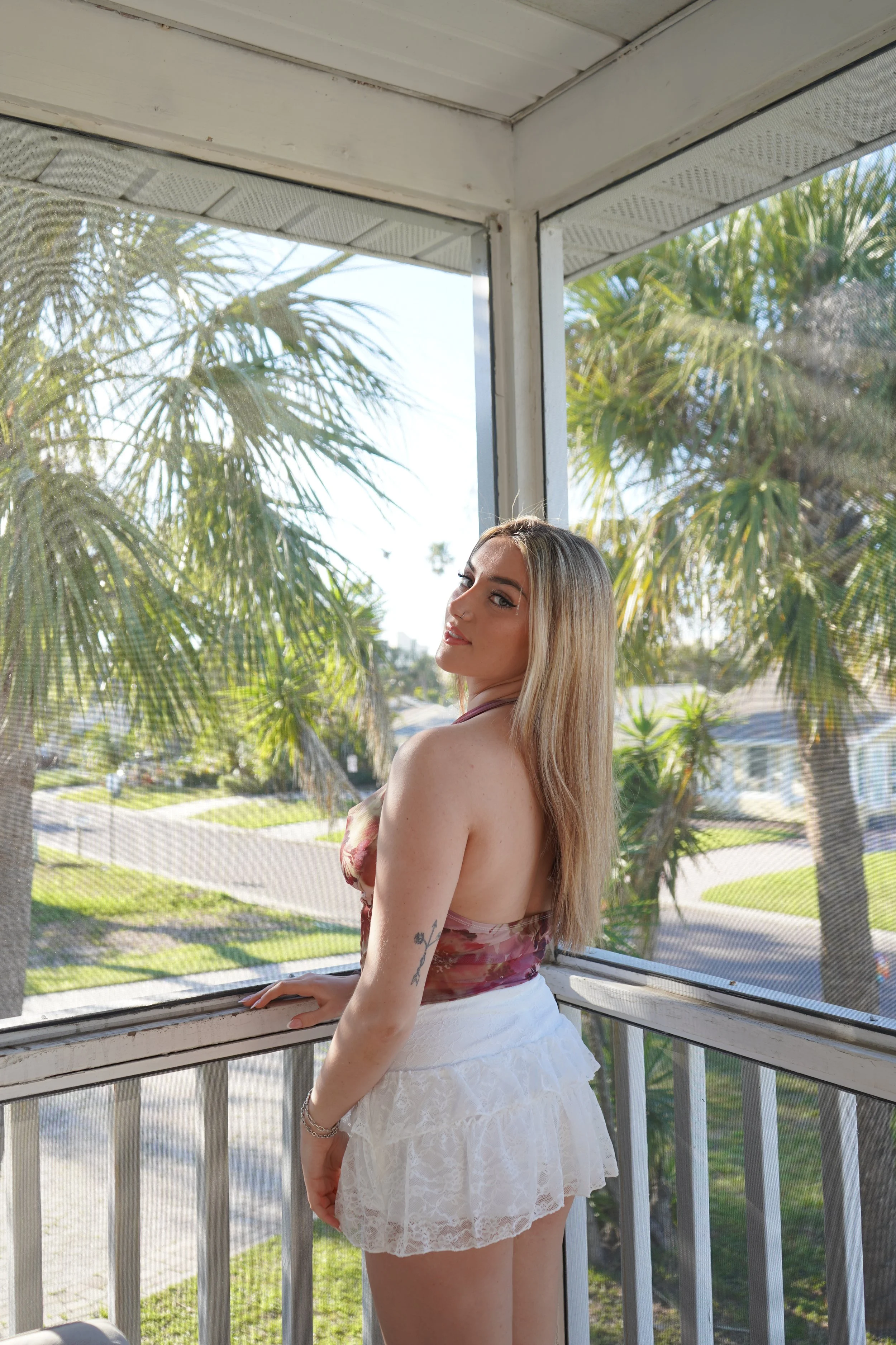 A woman with long blonde hair and tattoos on her arm, standing on a screened porch, looking over her shoulder, wearing a floral top and white lace skirt, with palm trees and a sunny neighborhood in the background.